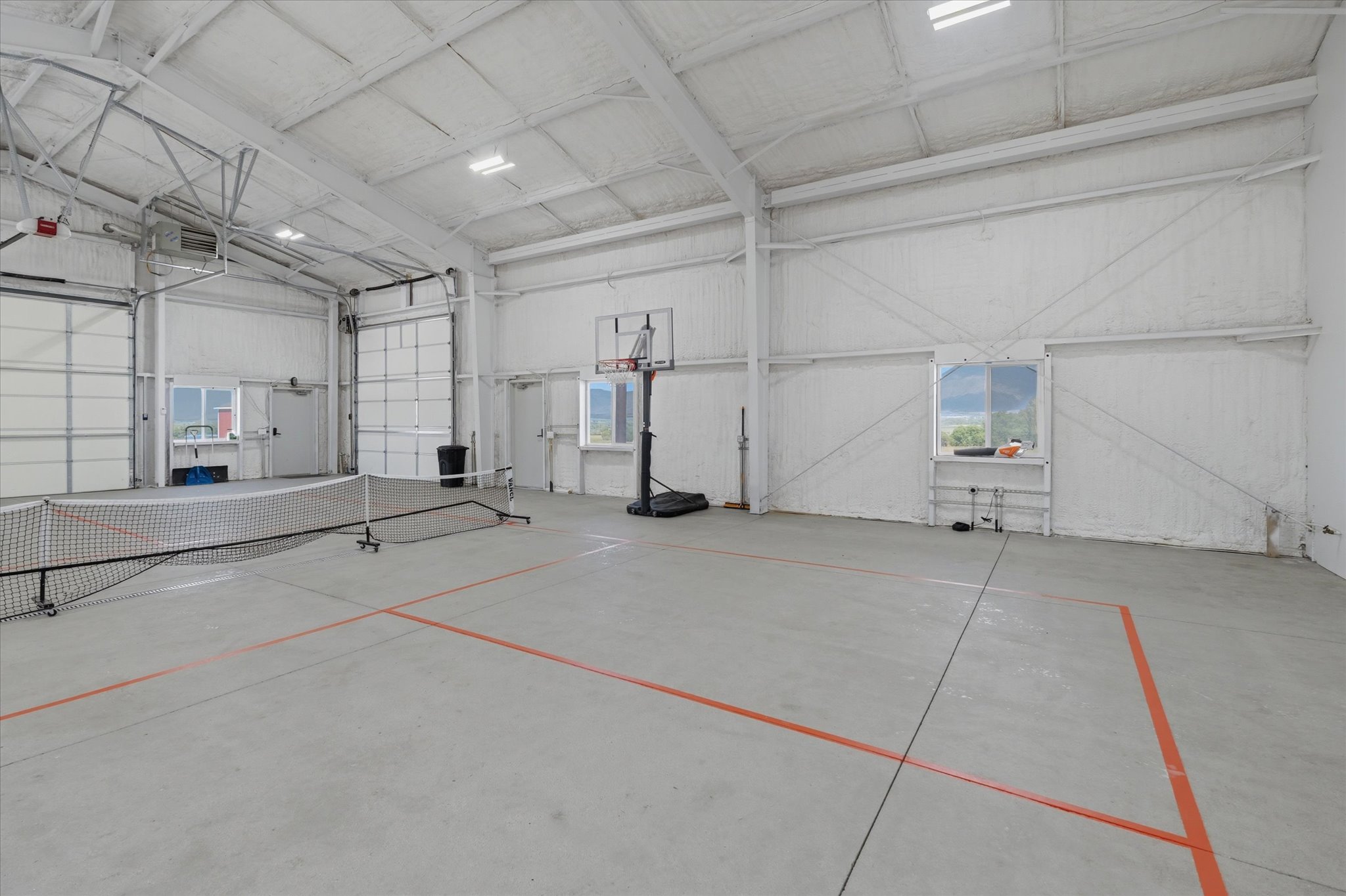 Spacious indoor court featuring concrete flooring with court lines, high ceilings, multiple roll-up doors, and fluorescent lighting