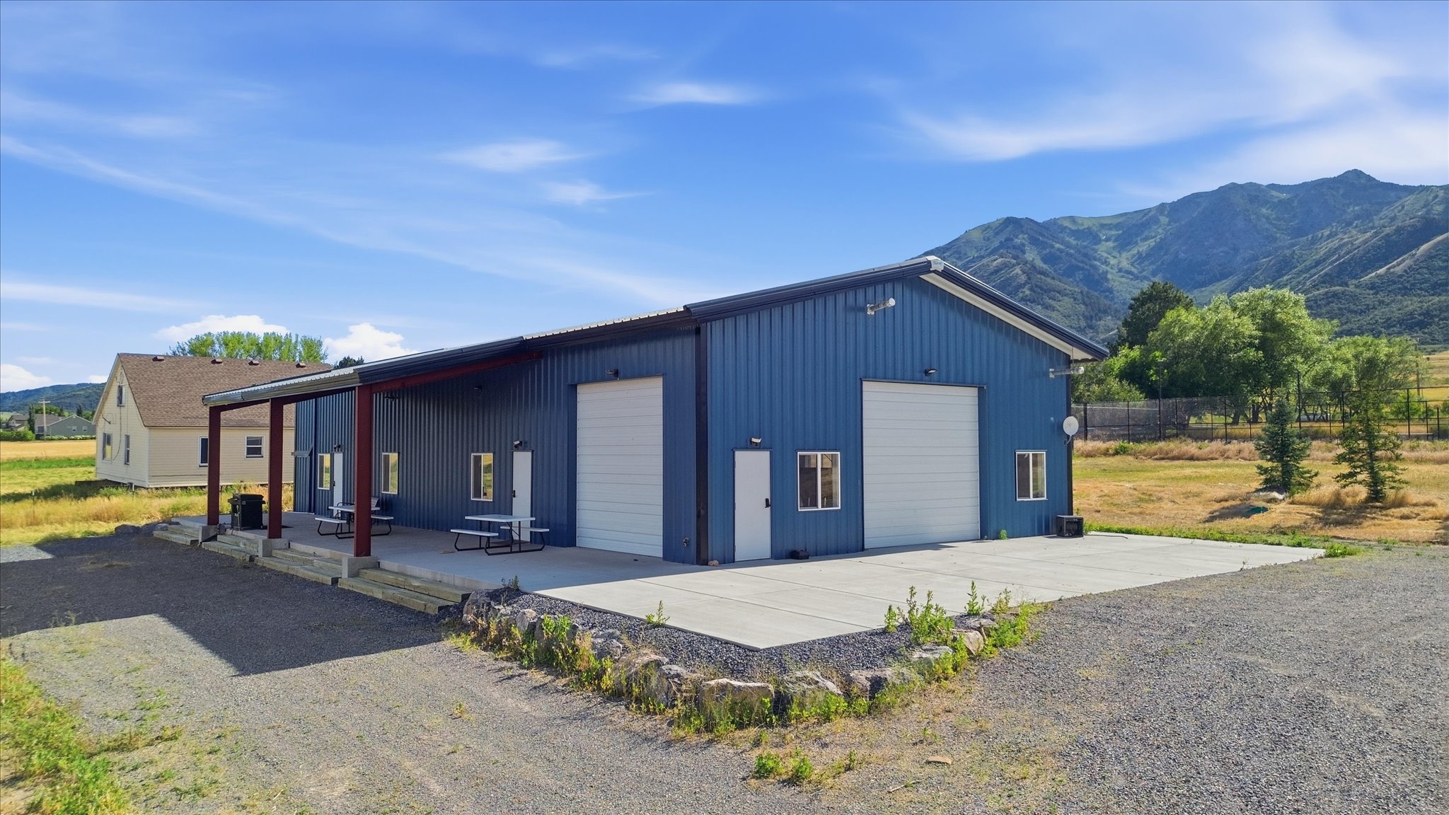 Blue metal-sided structure featuring two large white overhead doors, multiple windows, and a covered concrete patio supported by red beams