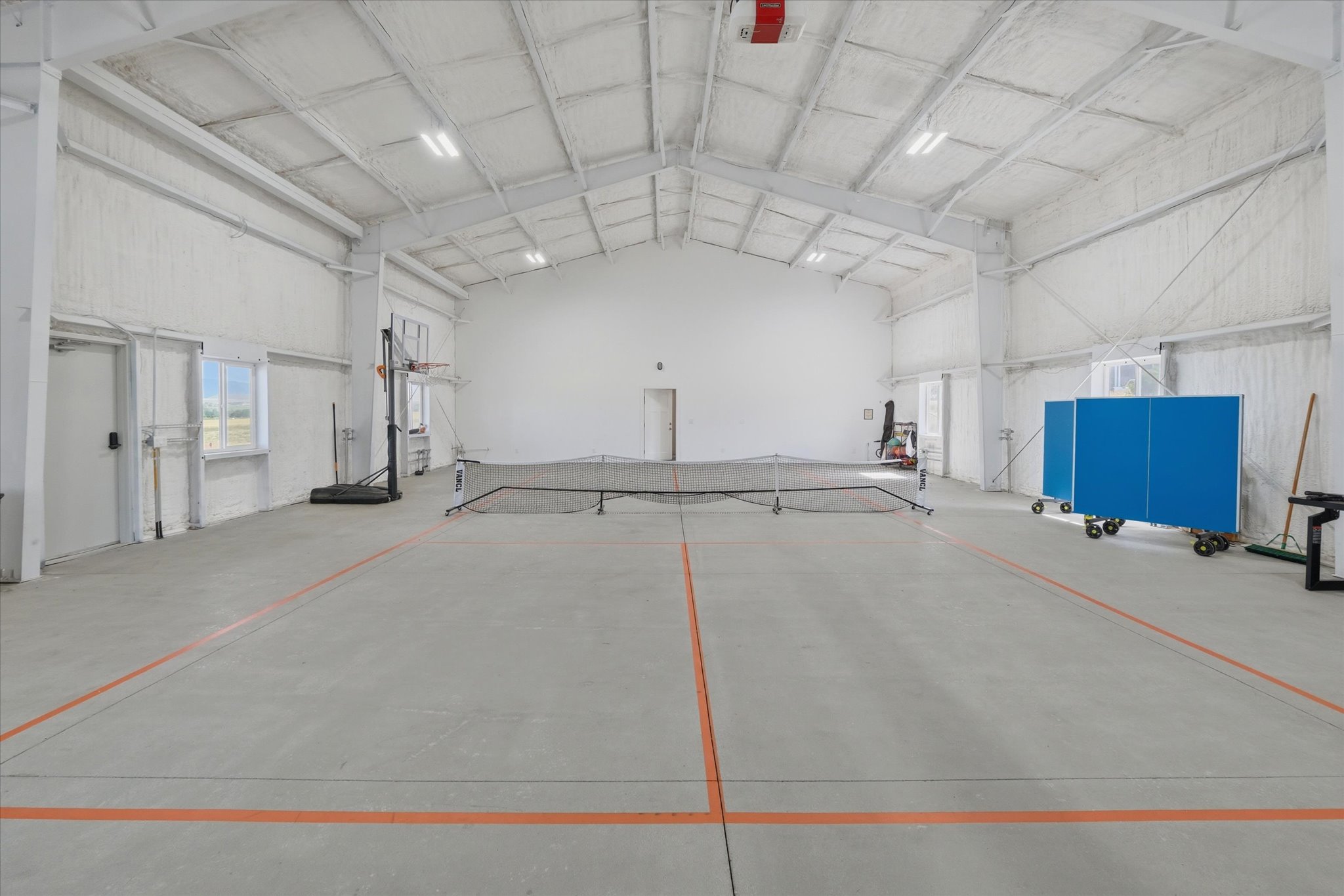 Spacious indoor court featuring concrete flooring with orange court lines, high ceilings with exposed trusses, and bright overhead lighting