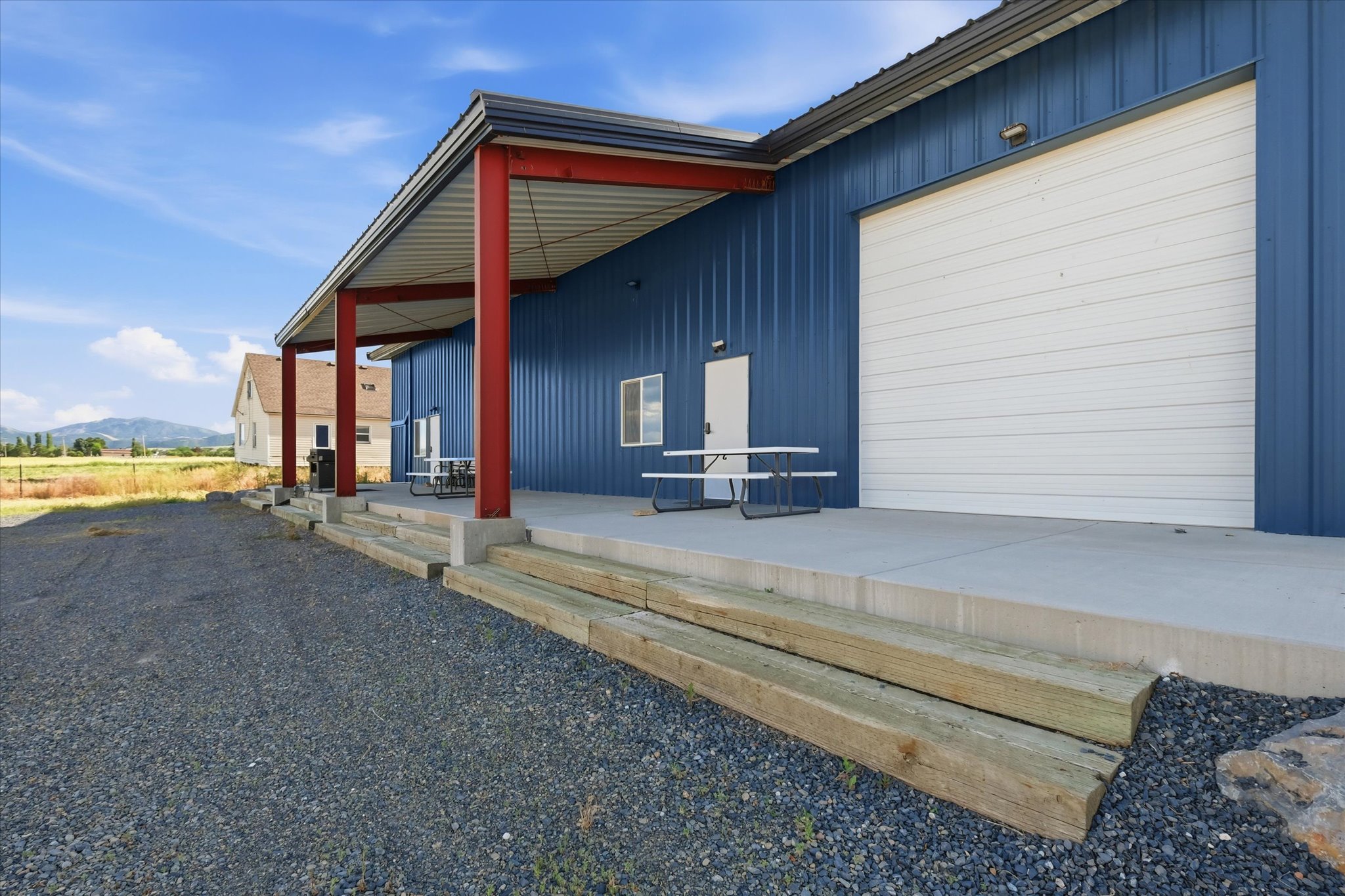 Blue corrugated metal siding building featuring a covered concrete porch with red support beams, two entry doors, and a large white roll-up door