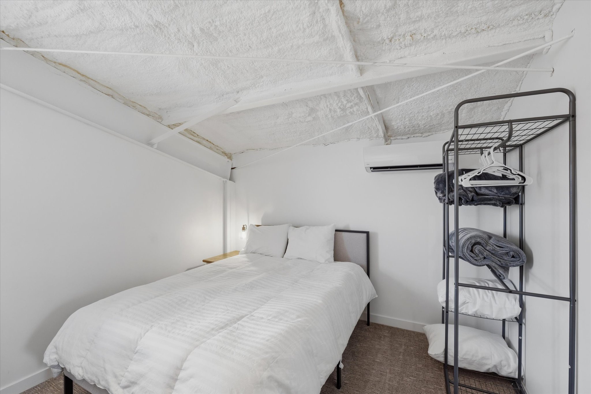 Bedroom featuring a wall-mounted AC unit, exposed beam ceiling with spray foam insulation, and a built-in wall sconce