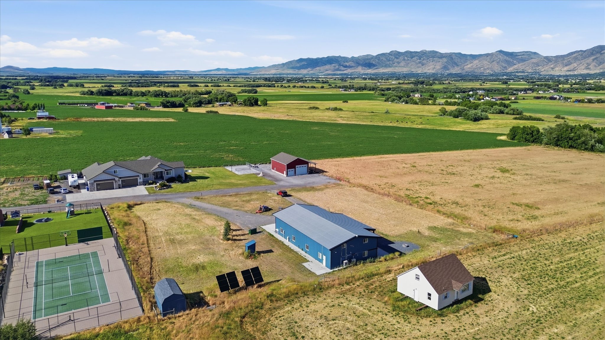 Expansive property featuring a modern ranch-style residence, a large blue industrial-style building, a red barn, a white outbuilding, and a fenced hardcourt