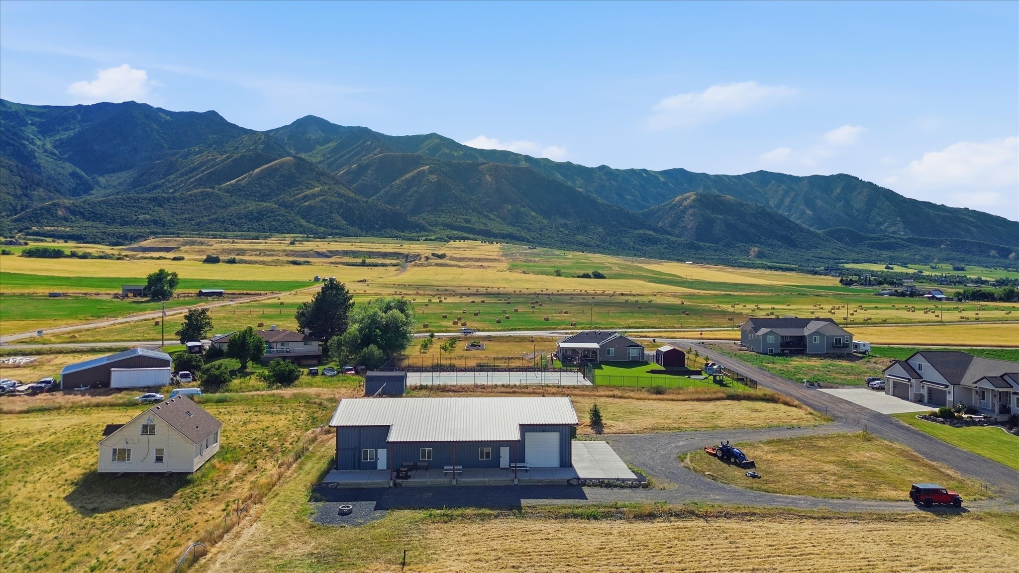 Expansive rural property featuring a large gray metal-sided structure with a white roll-up garage door, a paved driveway, a separate white residential structure, and a mountainous backdrop