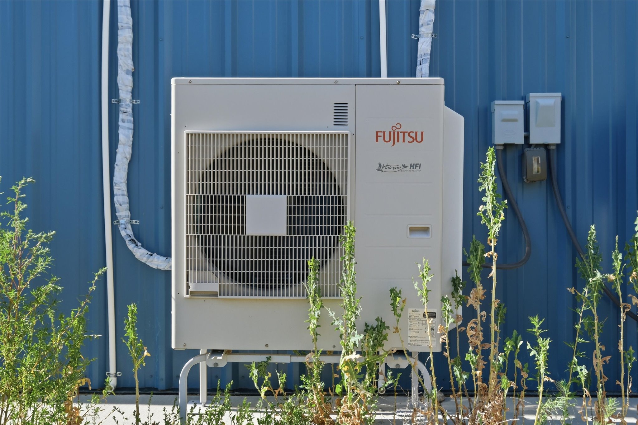 Exterior building infrastructure featuring a Fujitsu HVAC unit, blue corrugated metal siding, and exterior electrical boxes