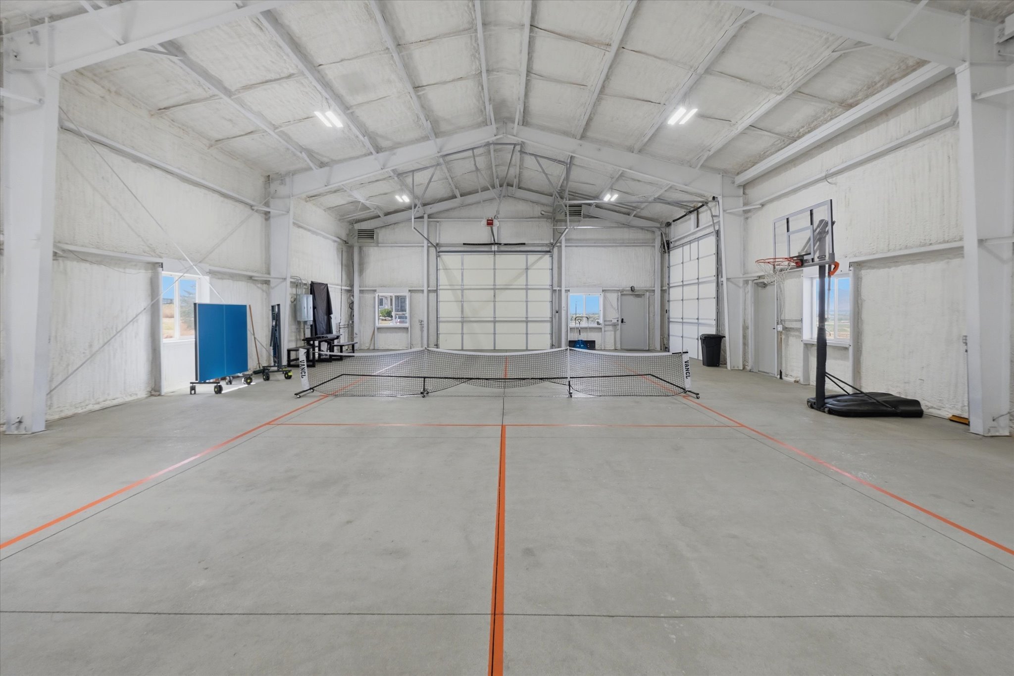 Spacious indoor recreational facility featuring concrete flooring with court line markings, high ceilings with exposed trusses, multiple overhead lighting fixtures, and large rollup doors