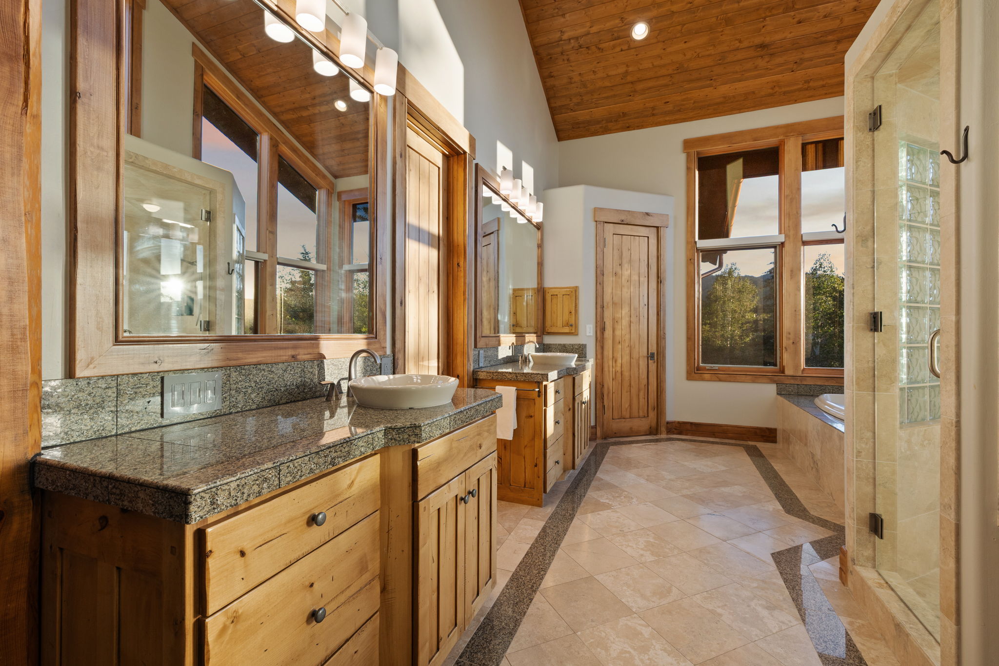 Spacious bathroom featuring natural wood cabinetry and trim, dual vanity with vessel sinks, granite countertops, tile flooring with dark accent border, and a glass-enclosed shower with glass block detailing