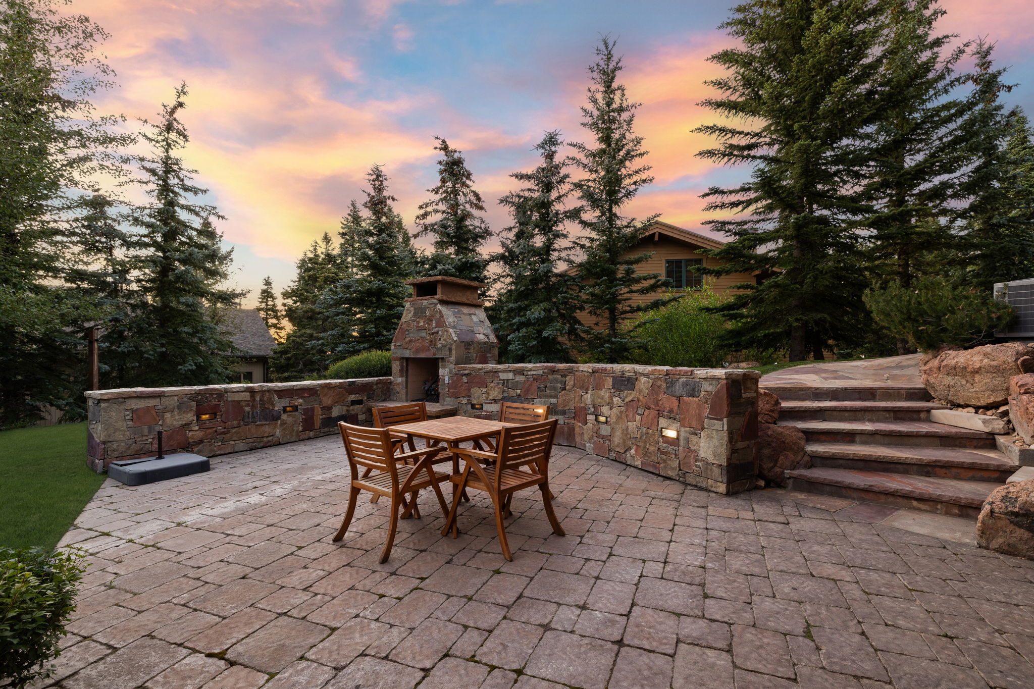 Expansive paver patio featuring a built-in stone fireplace, stone retaining walls with integrated lighting, and stone steps