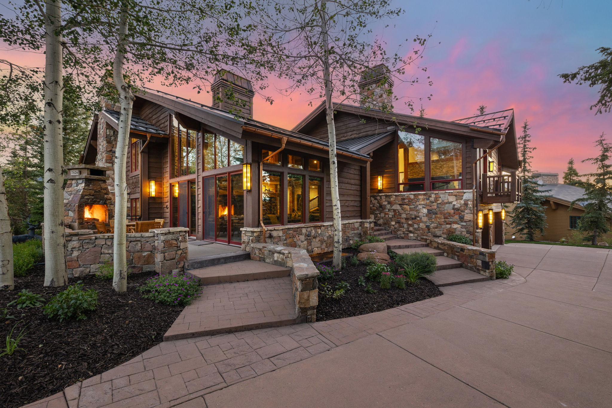 Stone and wood-sided exterior featuring multiple large windows, an outdoor fireplace, a stone retaining wall, and a stamped concrete driveway