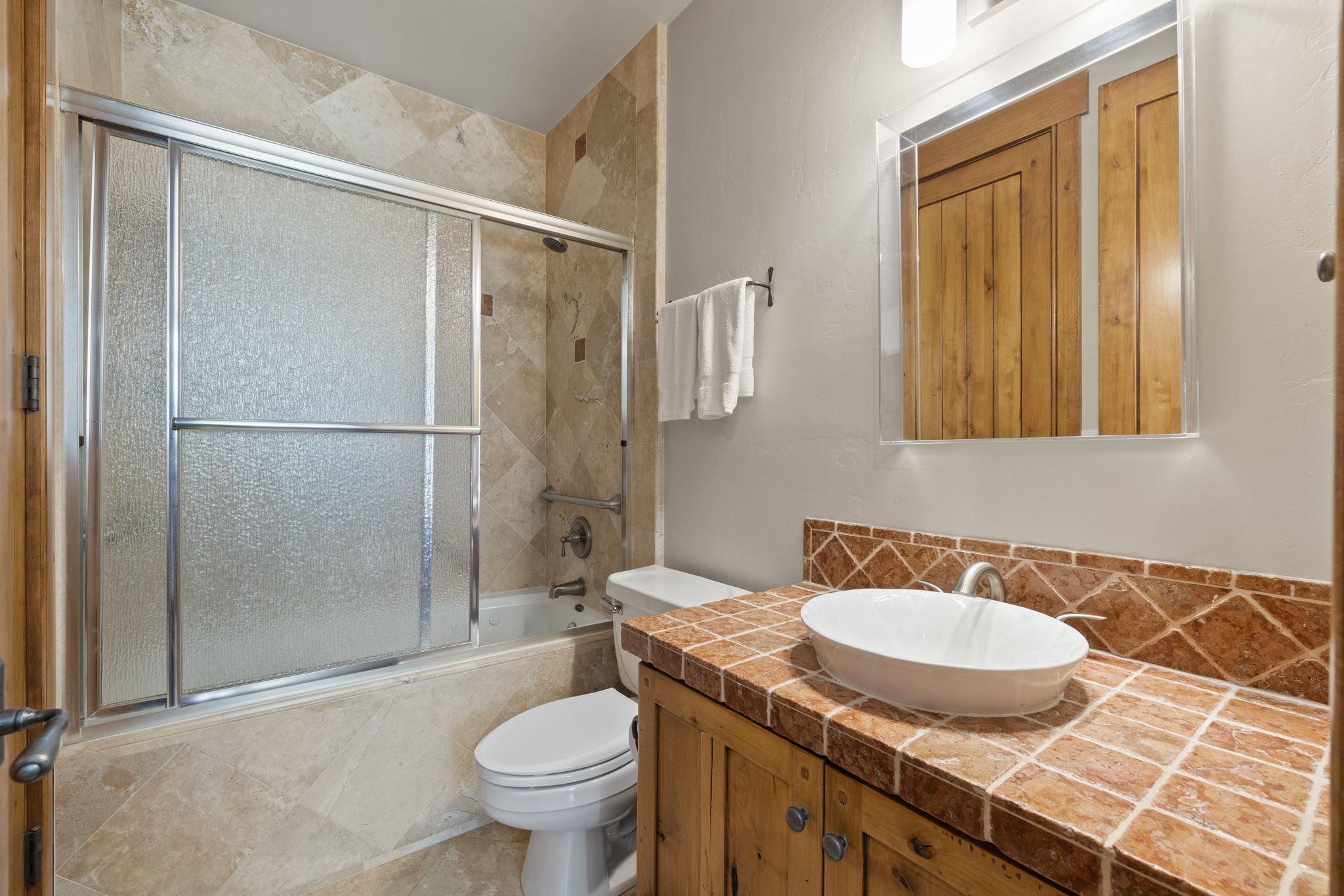 Bathroom featuring a tile-surround tub/shower with sliding glass doors, a white toilet, and a wood-finish vanity with a tiled countertop and vessel sink