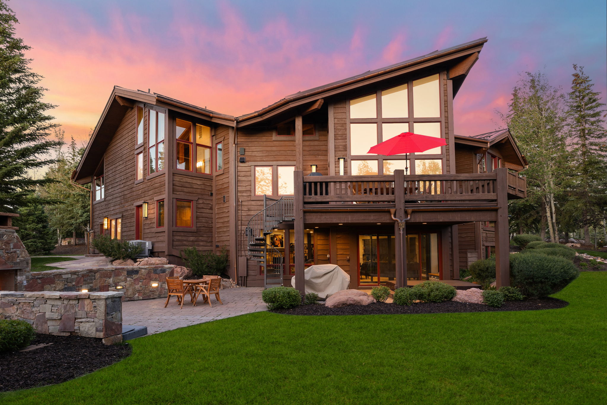 Multi-level wooden exterior with a stone patio, an upper-level deck, expansive windows, and a spiral staircase