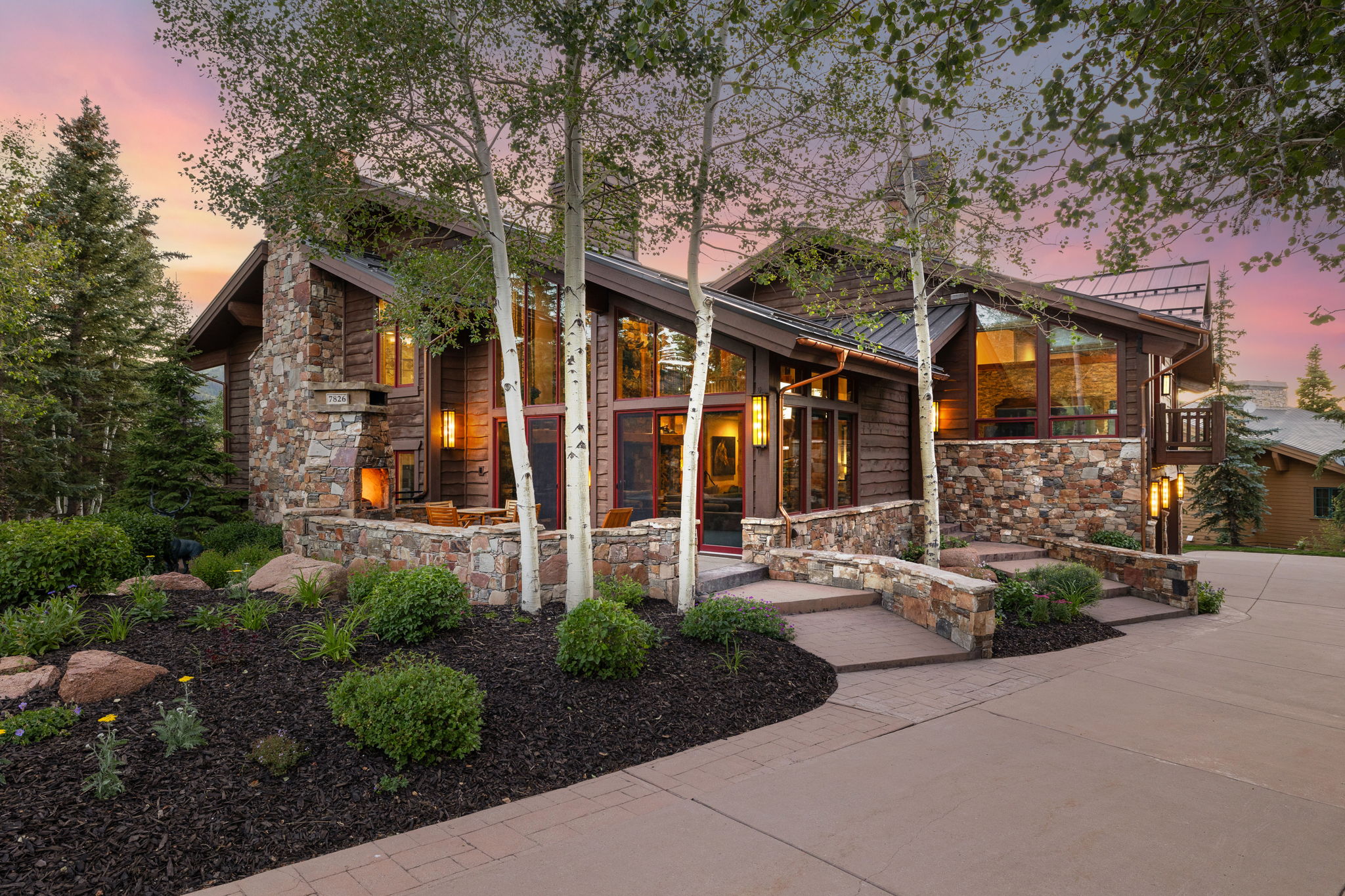 Rustic modern exterior featuring extensive stone masonry, natural wood siding, floor-to-ceiling windows, and integrated outdoor lighting