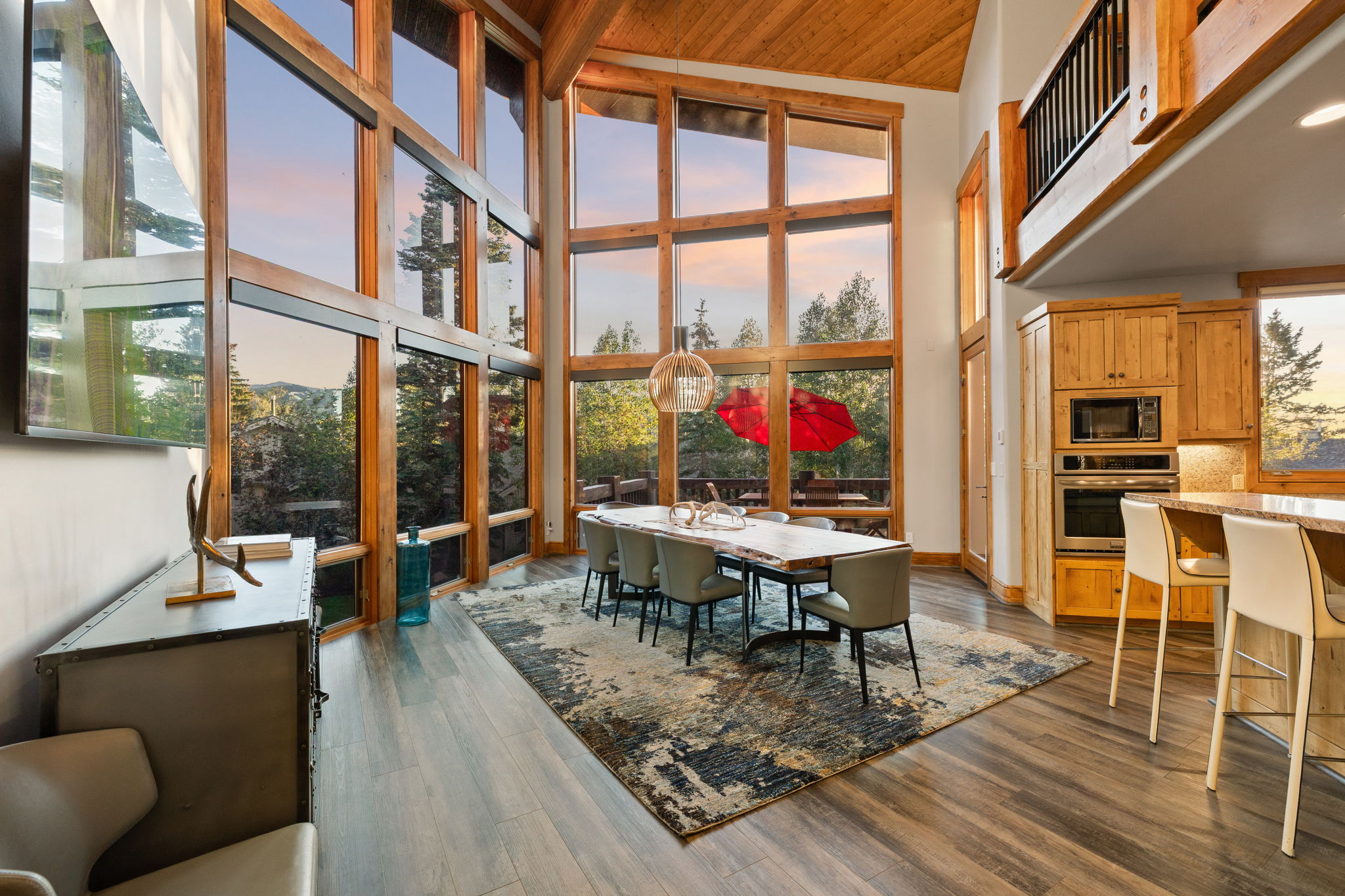 Vaulted ceiling with exposed wood beams, extensive floor-to-ceiling windows, wood-finish flooring, built-in stainless steel appliances, and a kitchen island with seating