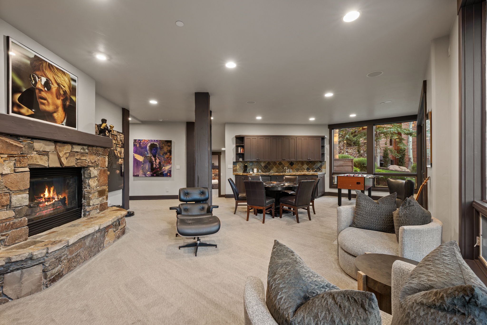 Expansive recreation space featuring a stone-clad fireplace with a hearth, recessed lighting, carpet flooring, a wet bar with dark cabinetry, and large windows