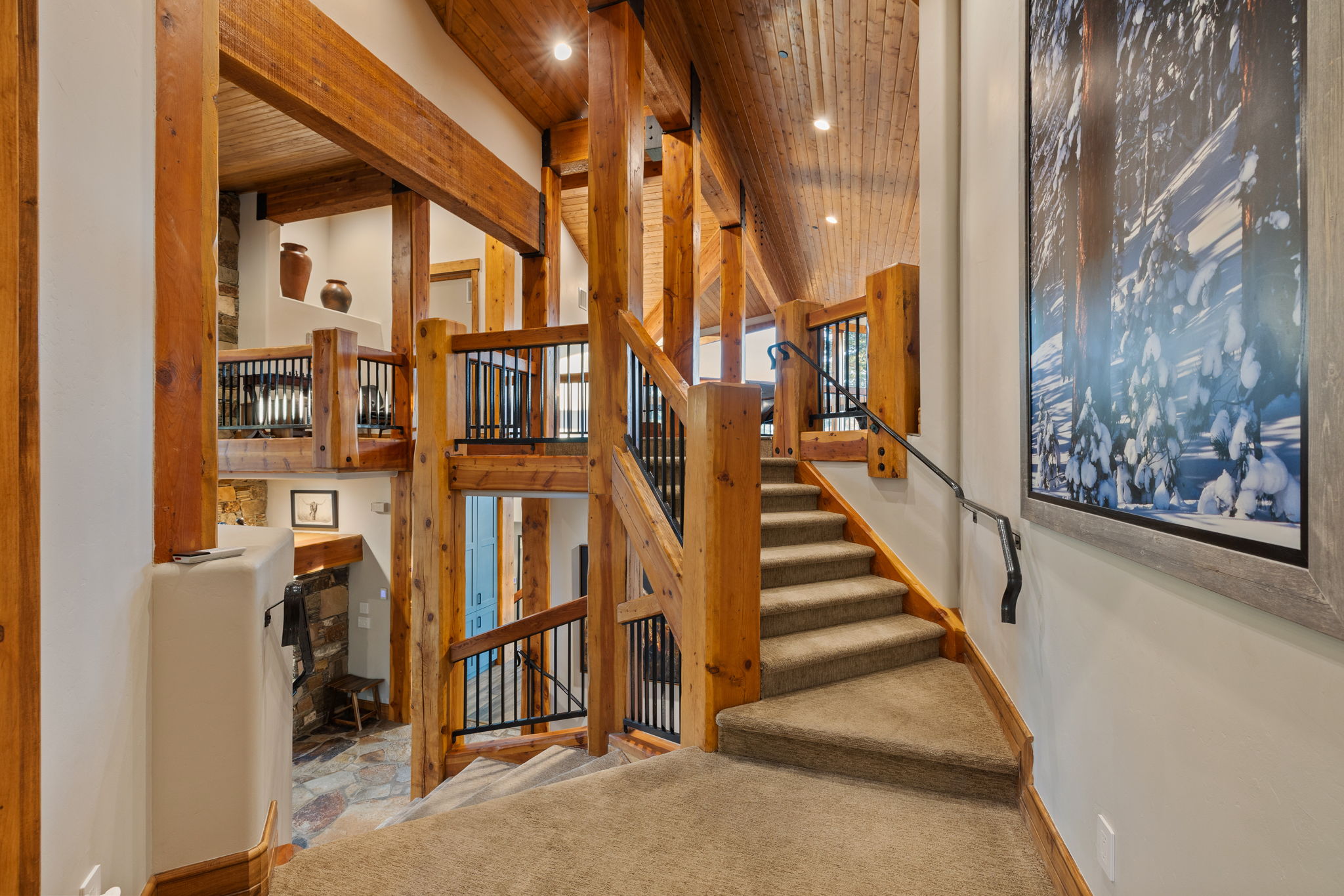 Multi-level interior featuring exposed timber beams, wood-finish vaulted ceilings, carpeted staircase with dark metal handrail, and a stone-clad fireplace