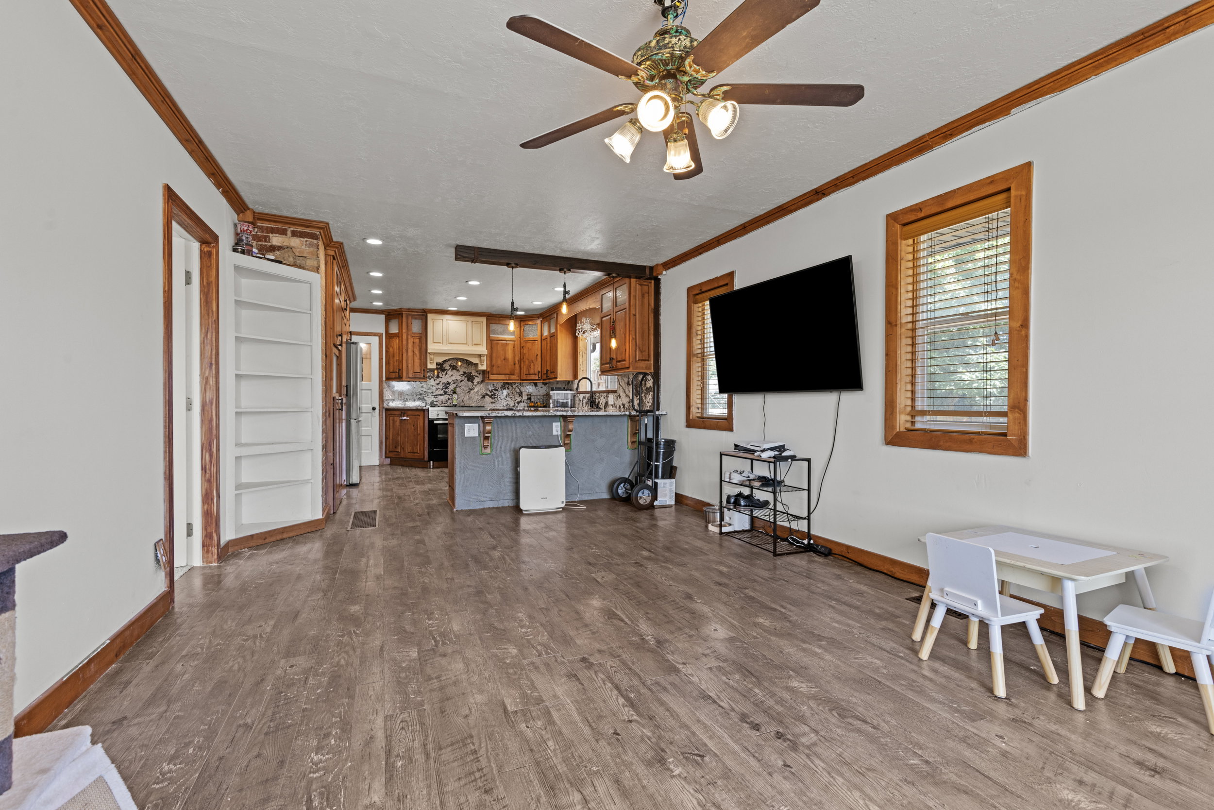 Expansive open-concept living area featuring wood-finish flooring, a ceiling fan with integrated lighting, and multiple windows with wood trim