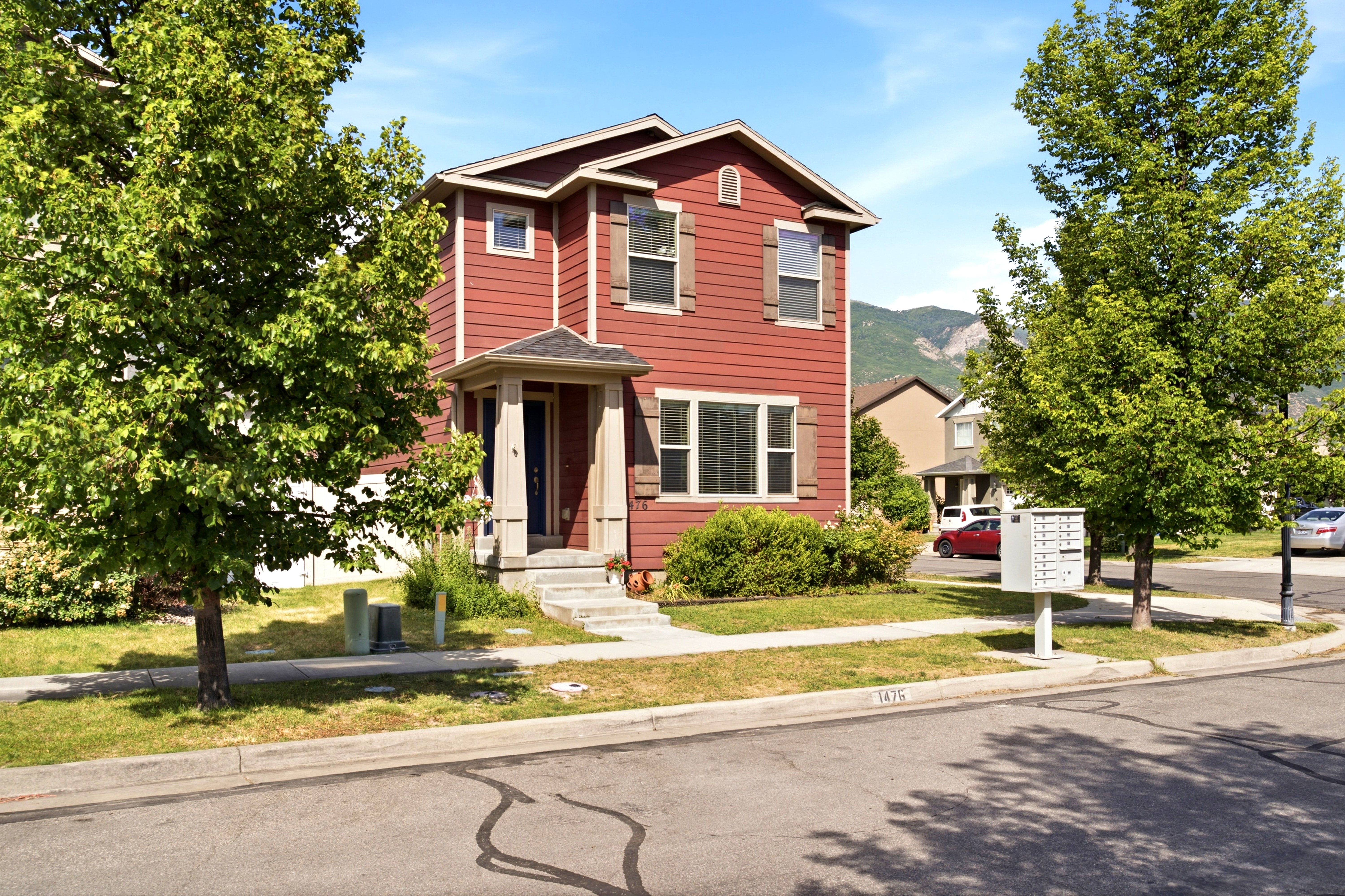 Two-story red siding residence featuring a covered front entry, contrasting window shutters, and established trees in the front yard