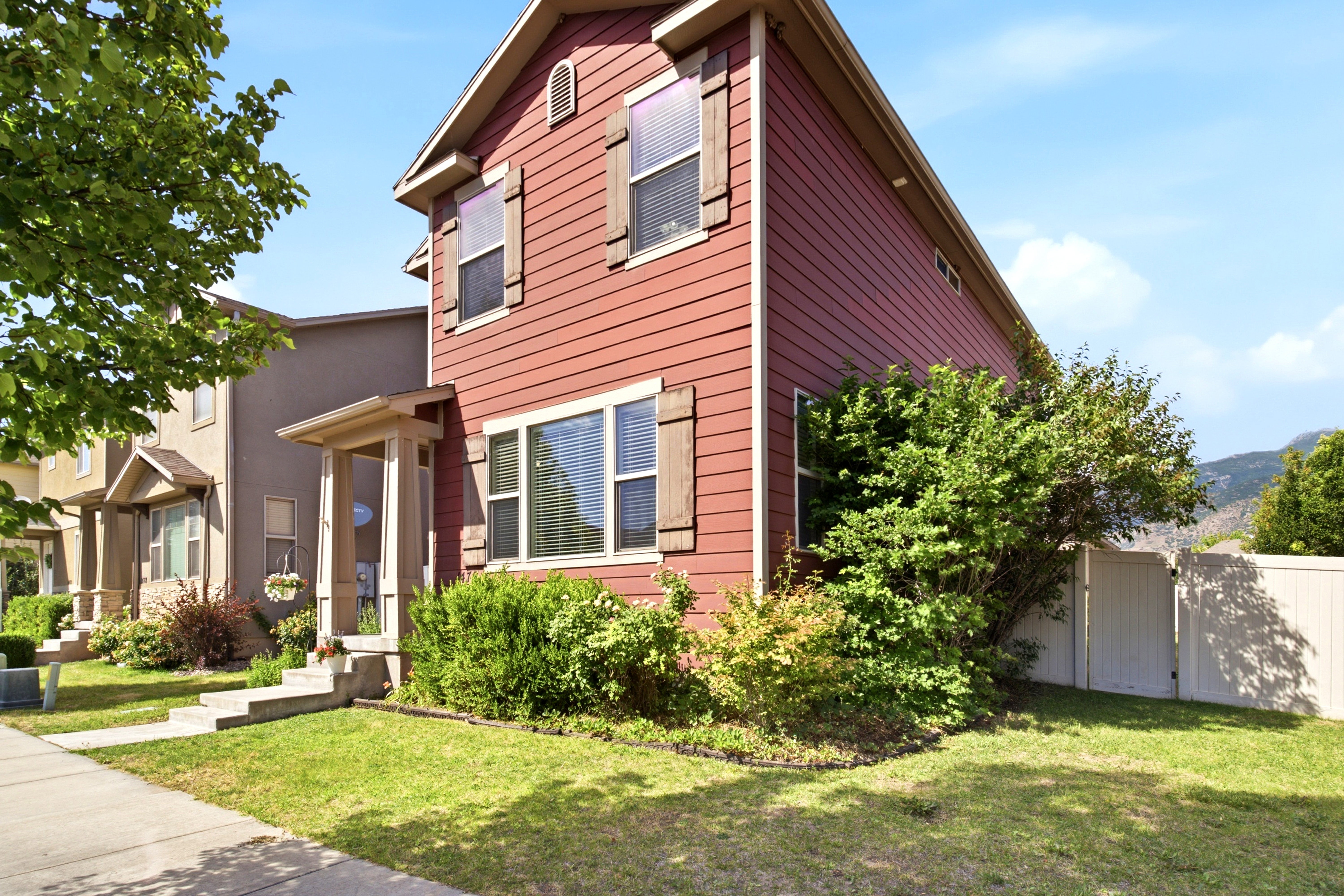 Red siding exterior with contrasting wood-finish shutters, a covered entry porch, established landscaping, and a white privacy fence