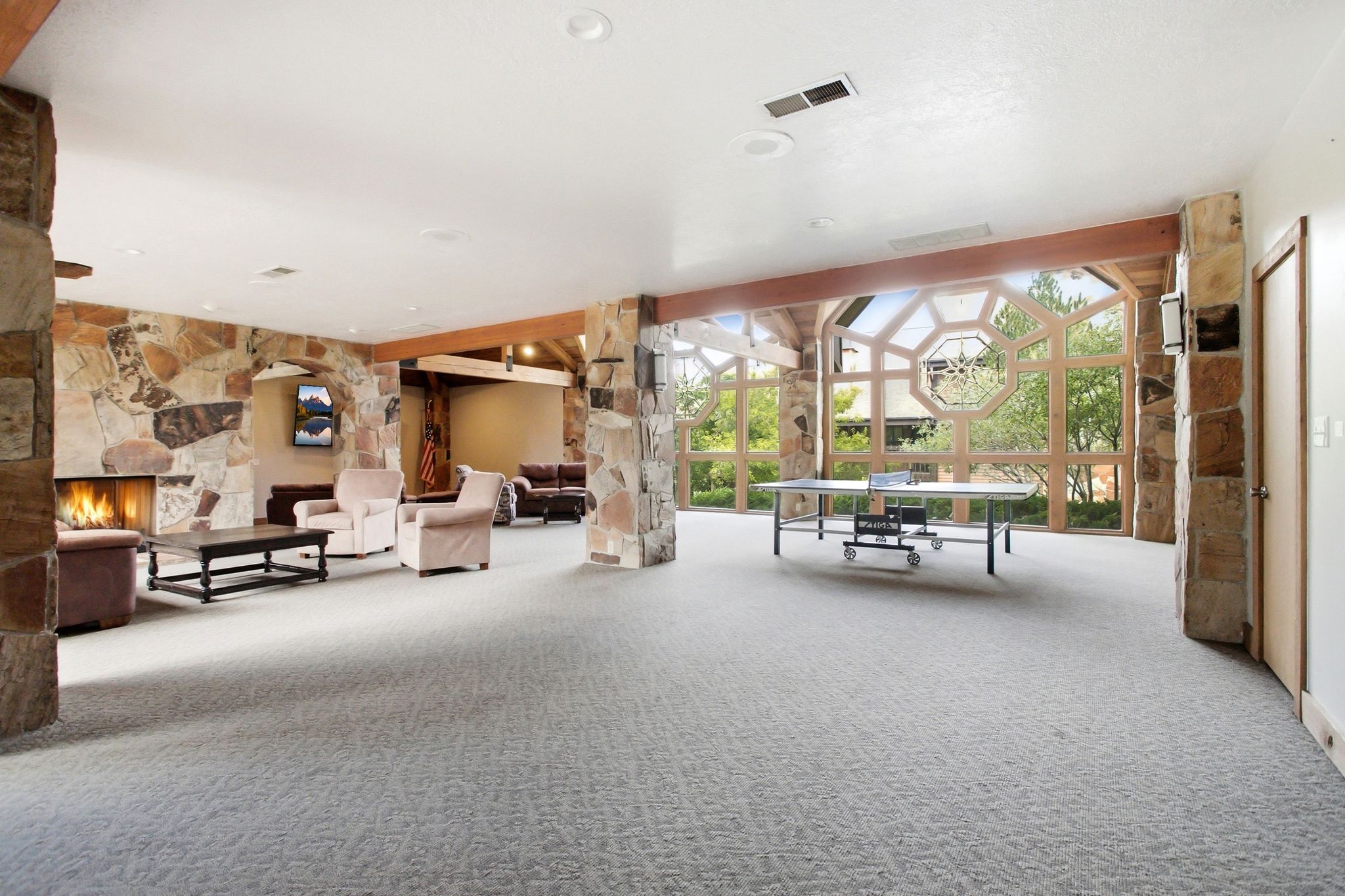 Spacious entertainment area featuring geometric window architecture, stone accent walls, exposed wood beams, recessed lighting, and soft carpet flooring
