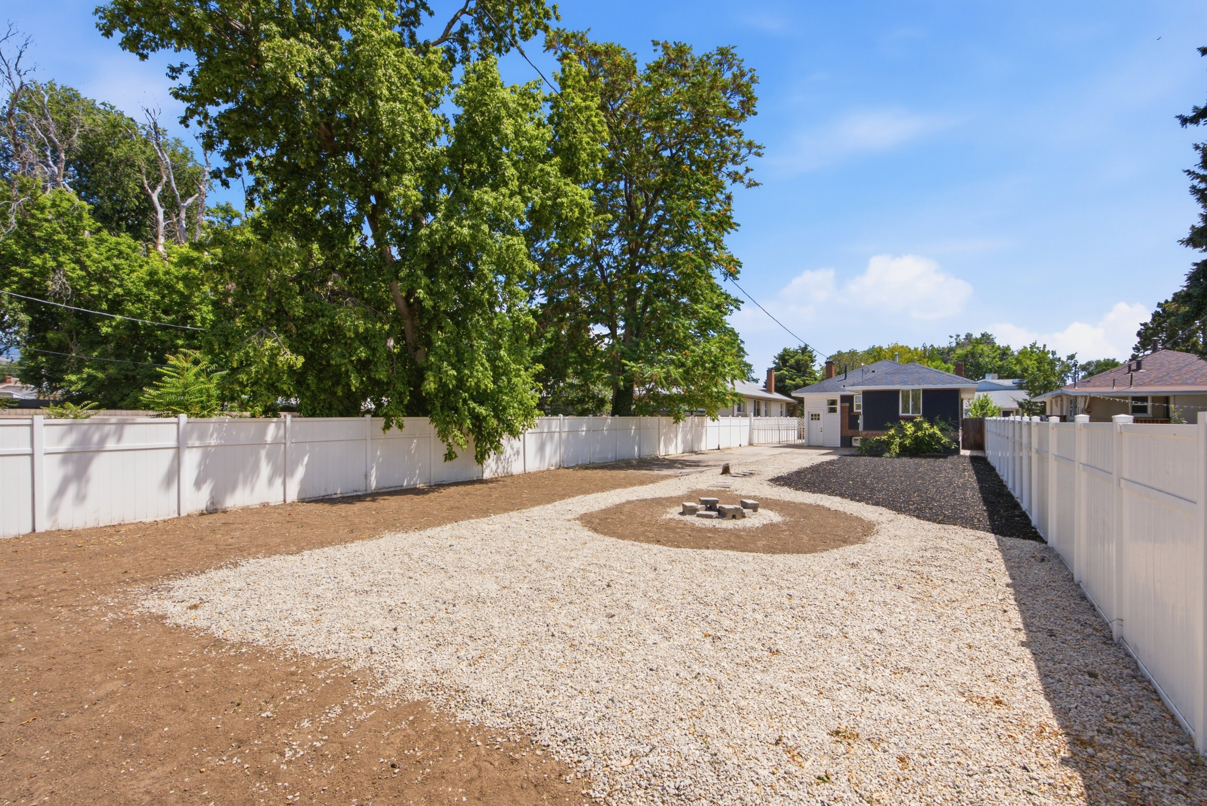 Spacious backyard featuring a white vinyl privacy fence, gravel landscaping, a central fire pit, and a dark-toned exterior residential structure