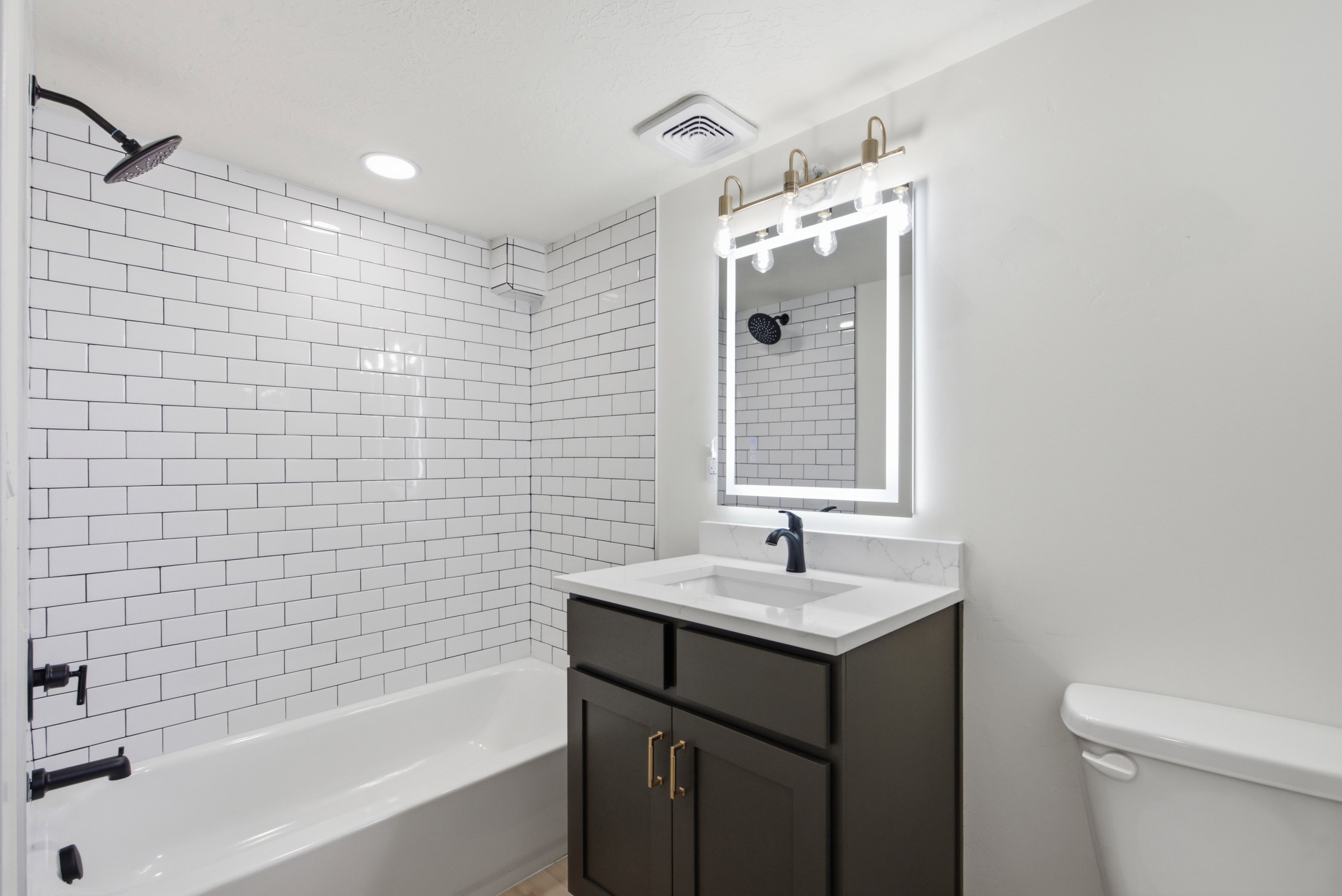 Bathroom featuring white subway tile surround, matte black shower fixtures, a dark wood-finish vanity with a white countertop and integrated sink, and an illuminated mirror with brass-finish lighting