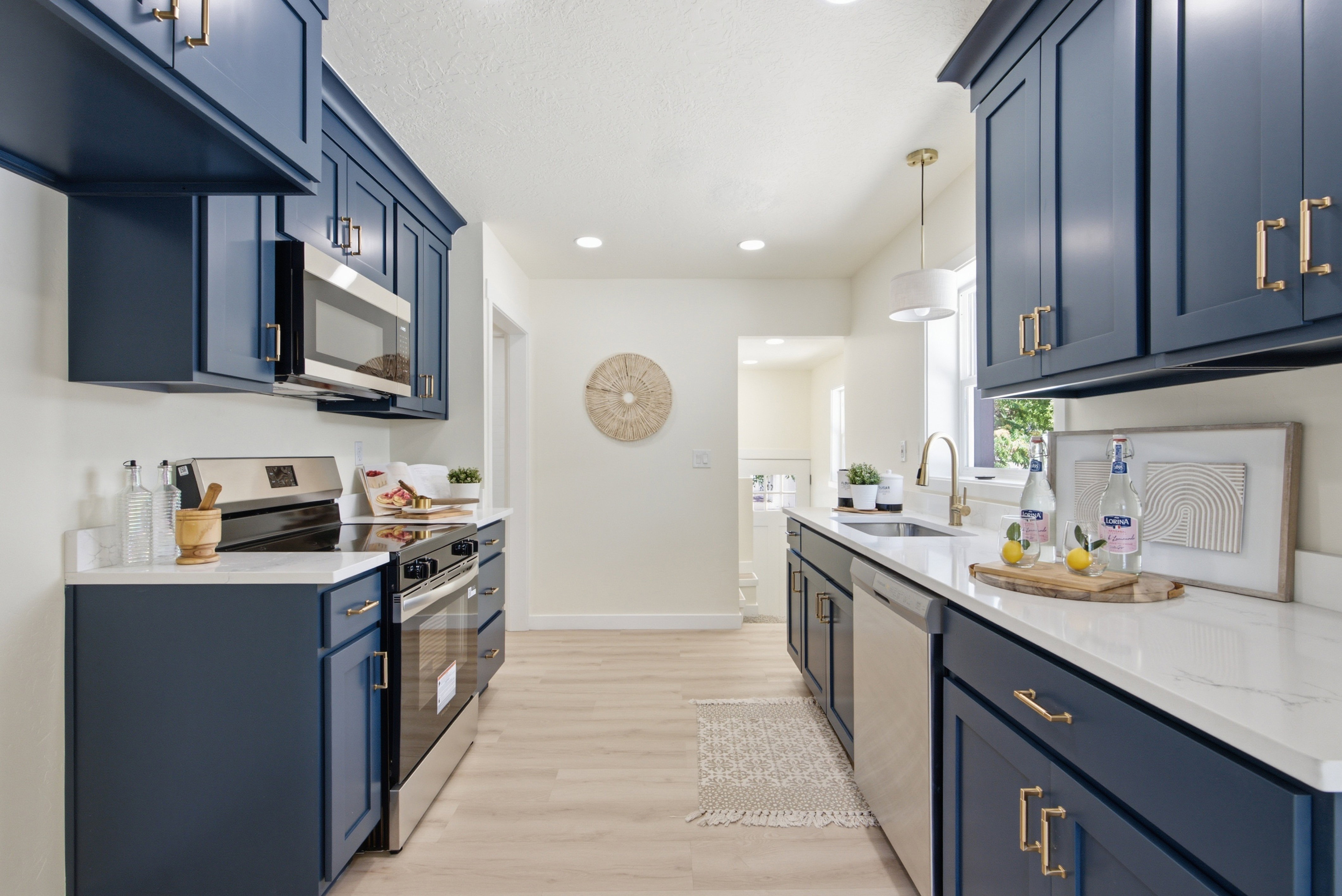 Modern kitchen featuring dark blue cabinetry with gold-tone hardware, stainless steel appliances, light wood-finish flooring, and white countertops