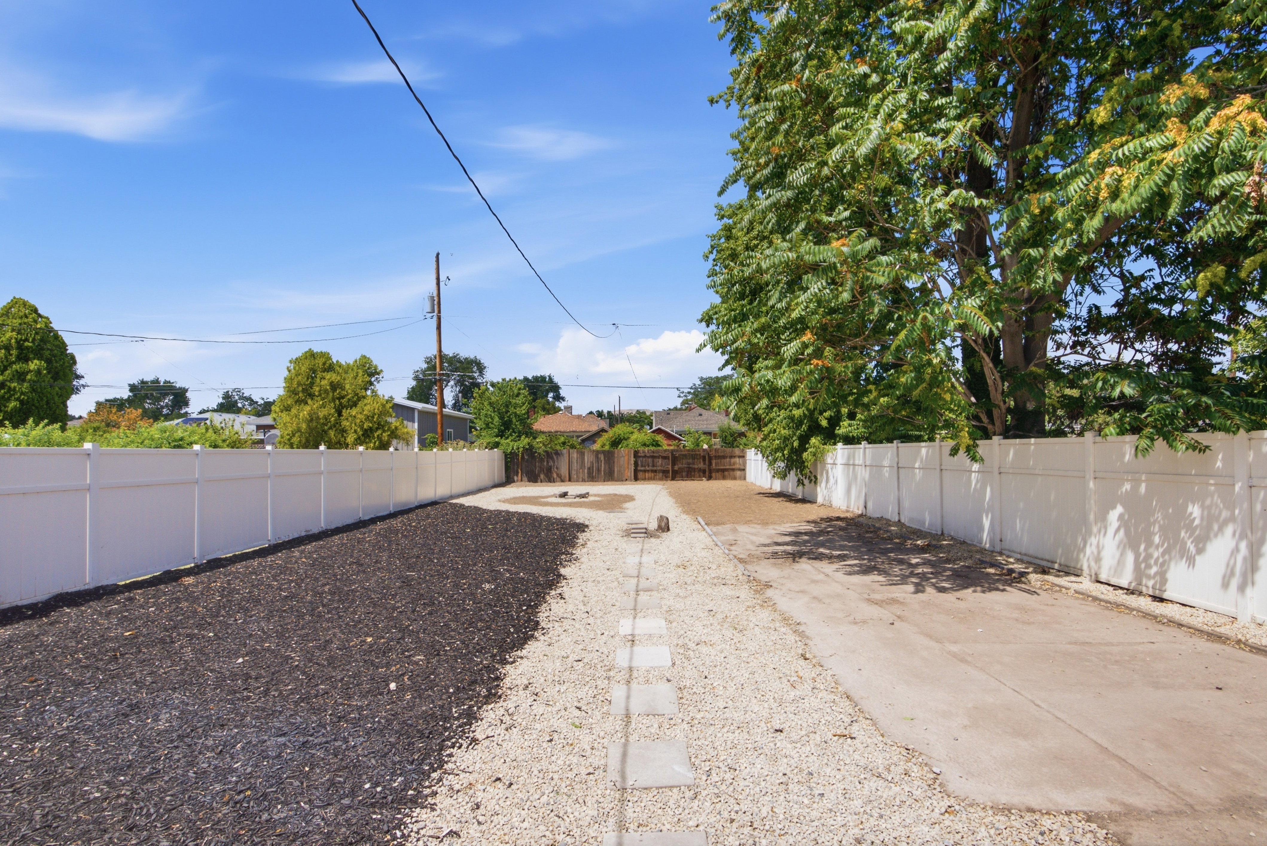 Spacious outdoor area featuring a white privacy fence, gravel and mulch landscaping, and mature trees