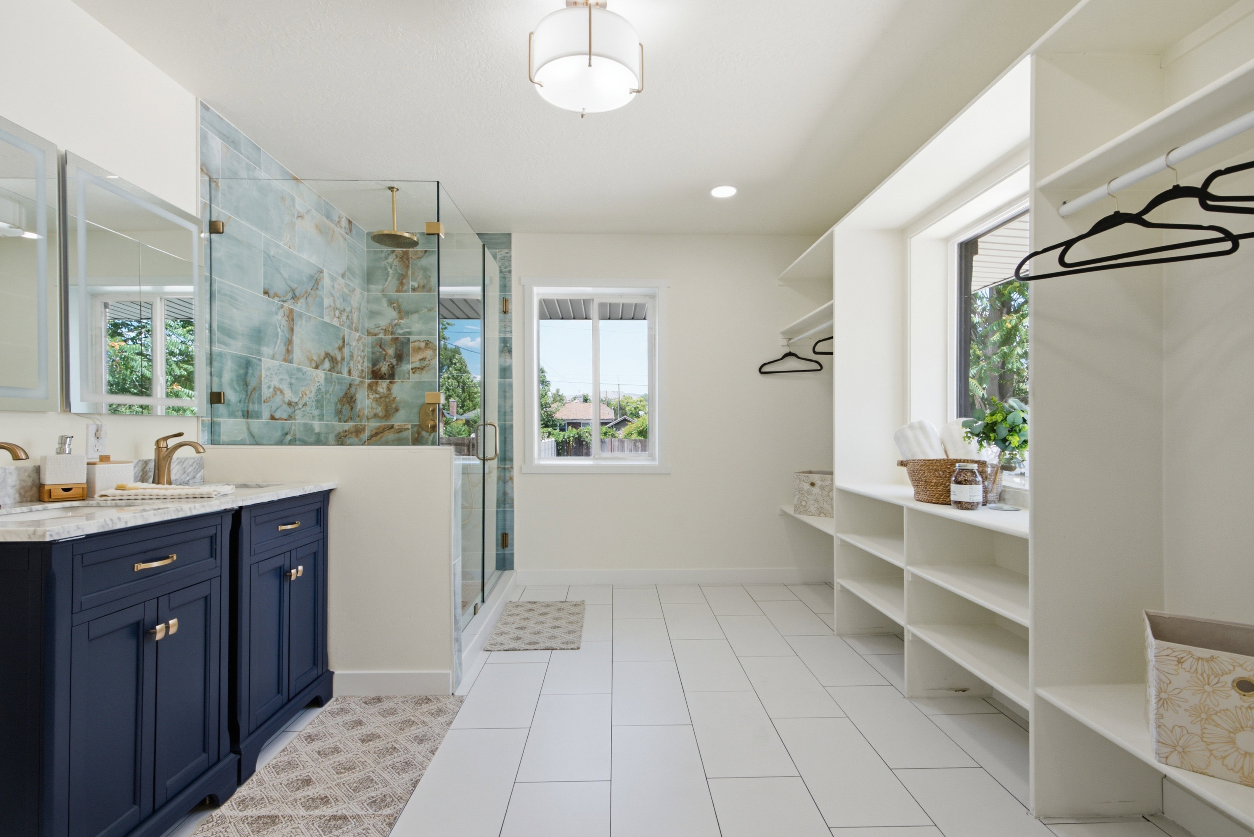 Expansive bathroom featuring a double vanity with navy cabinetry, a glass-enclosed shower with decorative tile, white tile flooring, and integrated open shelving and hanging storage