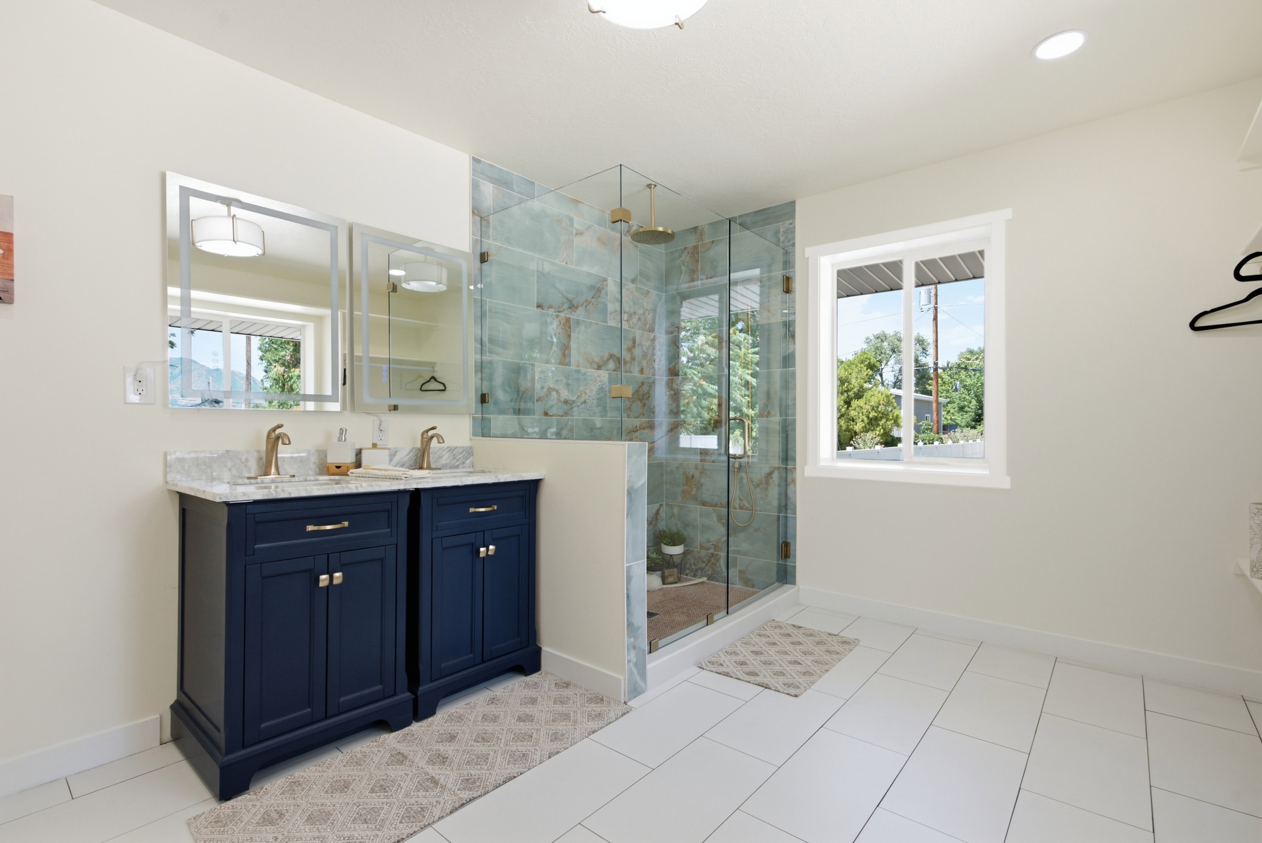 Spacious bathroom featuring a dual vanity with navy cabinetry and gold-toned fixtures, a glass-enclosed shower with decorative wall tiling, and white rectangular floor tiles