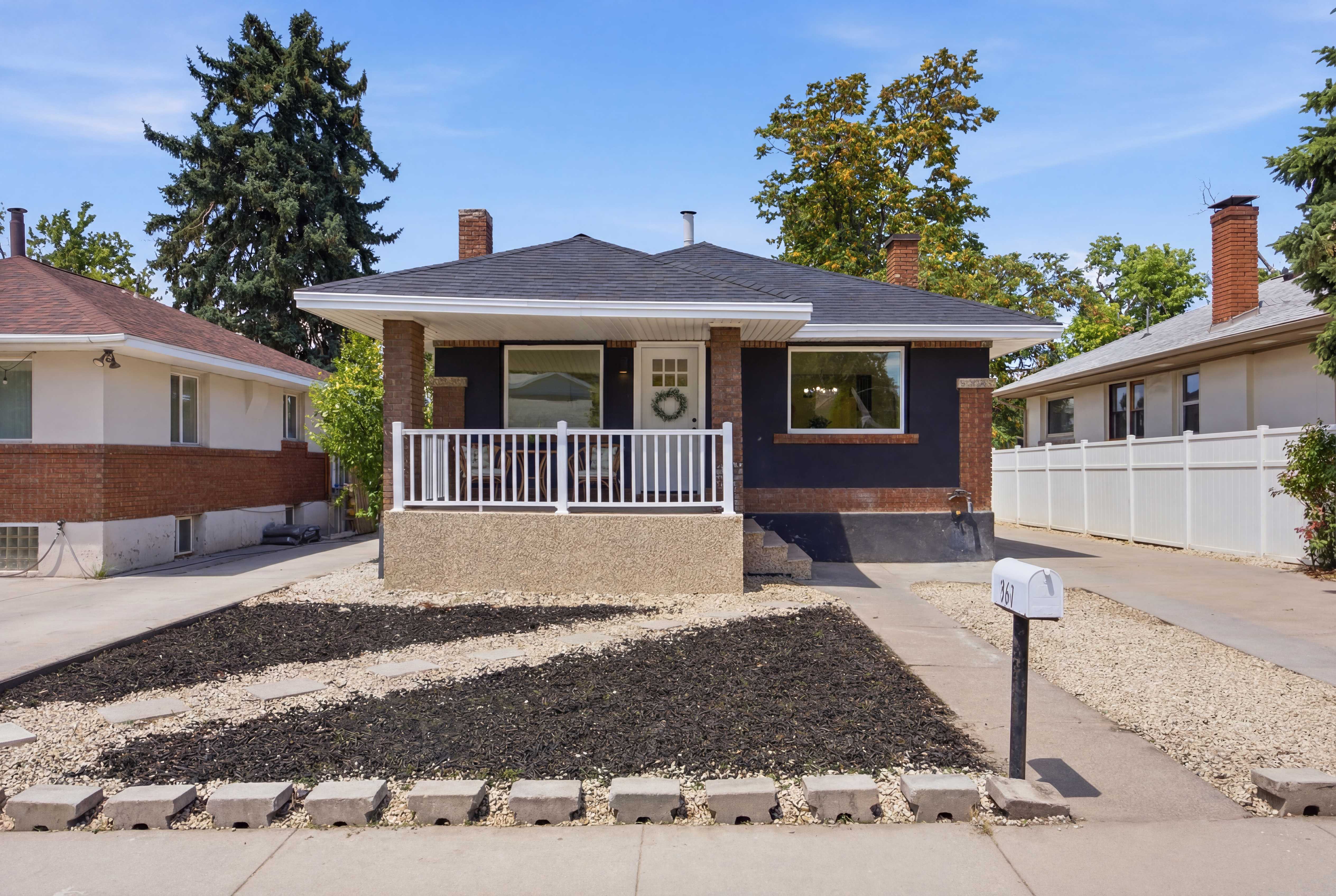 Contemporary bungalow featuring a covered front porch with white railing, a dark exterior finish with brick accents, and a shingle roof