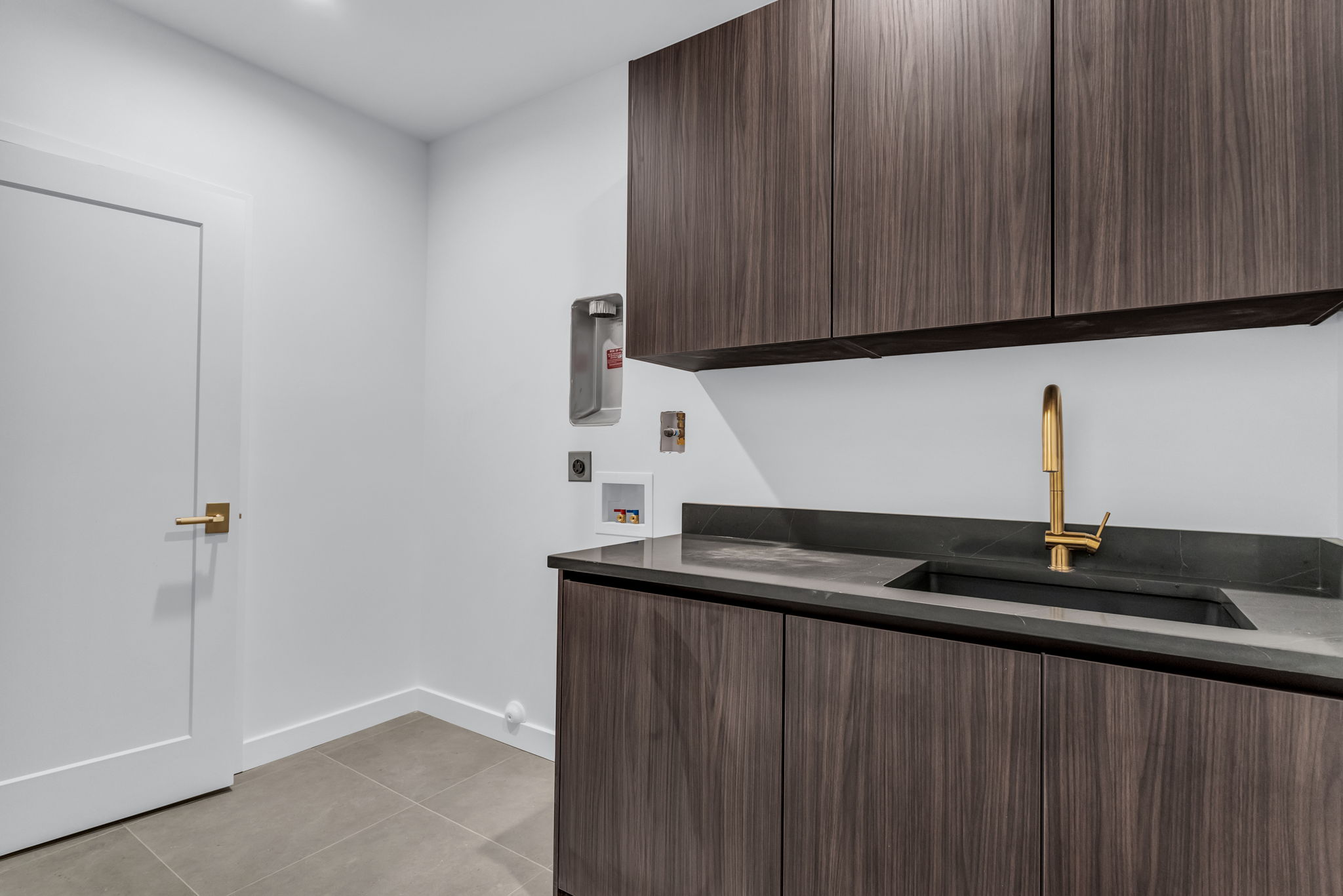 Laundry space featuring wood-finish cabinetry, a dark countertop, an undermount sink with a brass-finish faucet, and light tile flooring