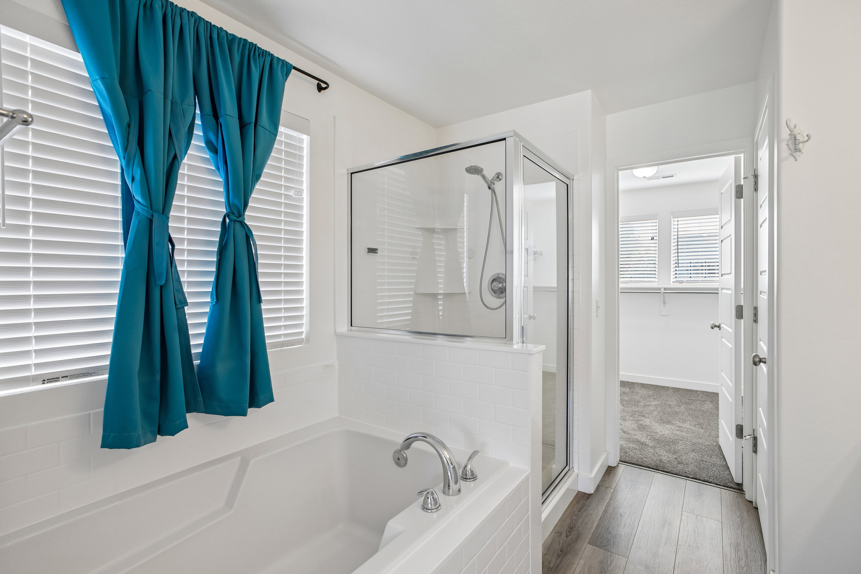 Soaking tub with chrome fixtures, glass-enclosed shower, white tile surround, and wood-finish flooring