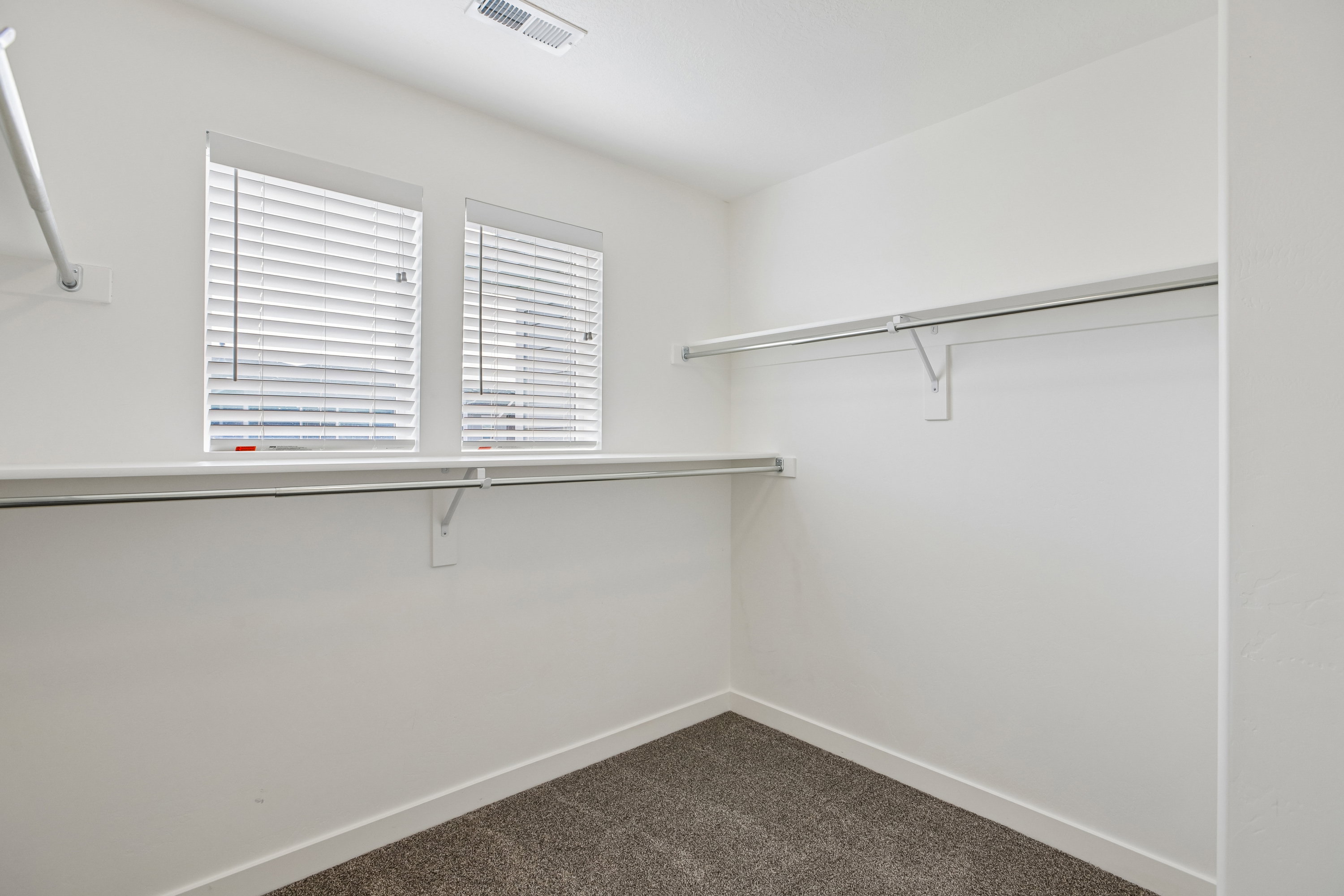 Spacious closet featuring two windows with horizontal blinds, overhead ventilation, white walls, and gray carpet flooring