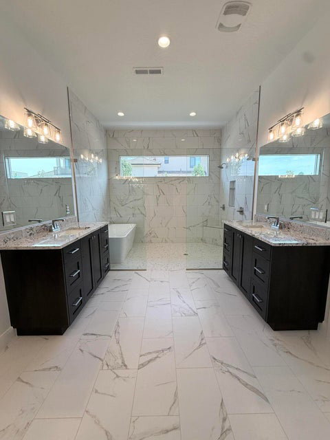Spacious bathroom featuring marble-look tile flooring, dual dark wood-finish vanities with light stone countertops, and a large walk-in shower with a freestanding tub