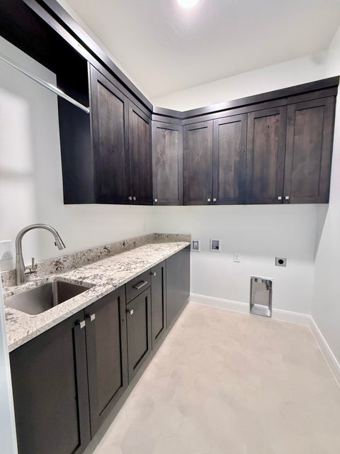 Laundry room featuring dark wood-finish cabinetry, light-toned countertops, a stainless steel sink with a brushed nickel faucet, and light-colored flooring