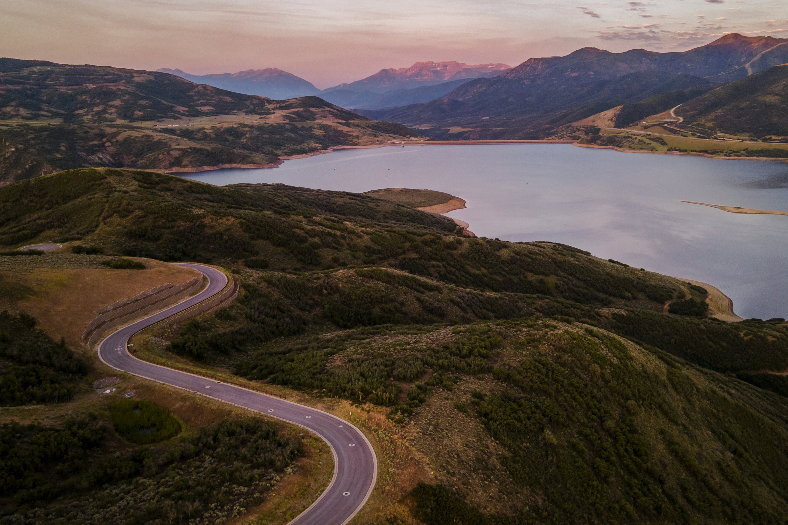 Winding paved road traversing a mountainous landscape with a large body of water, distant peaks, and varied natural terrain