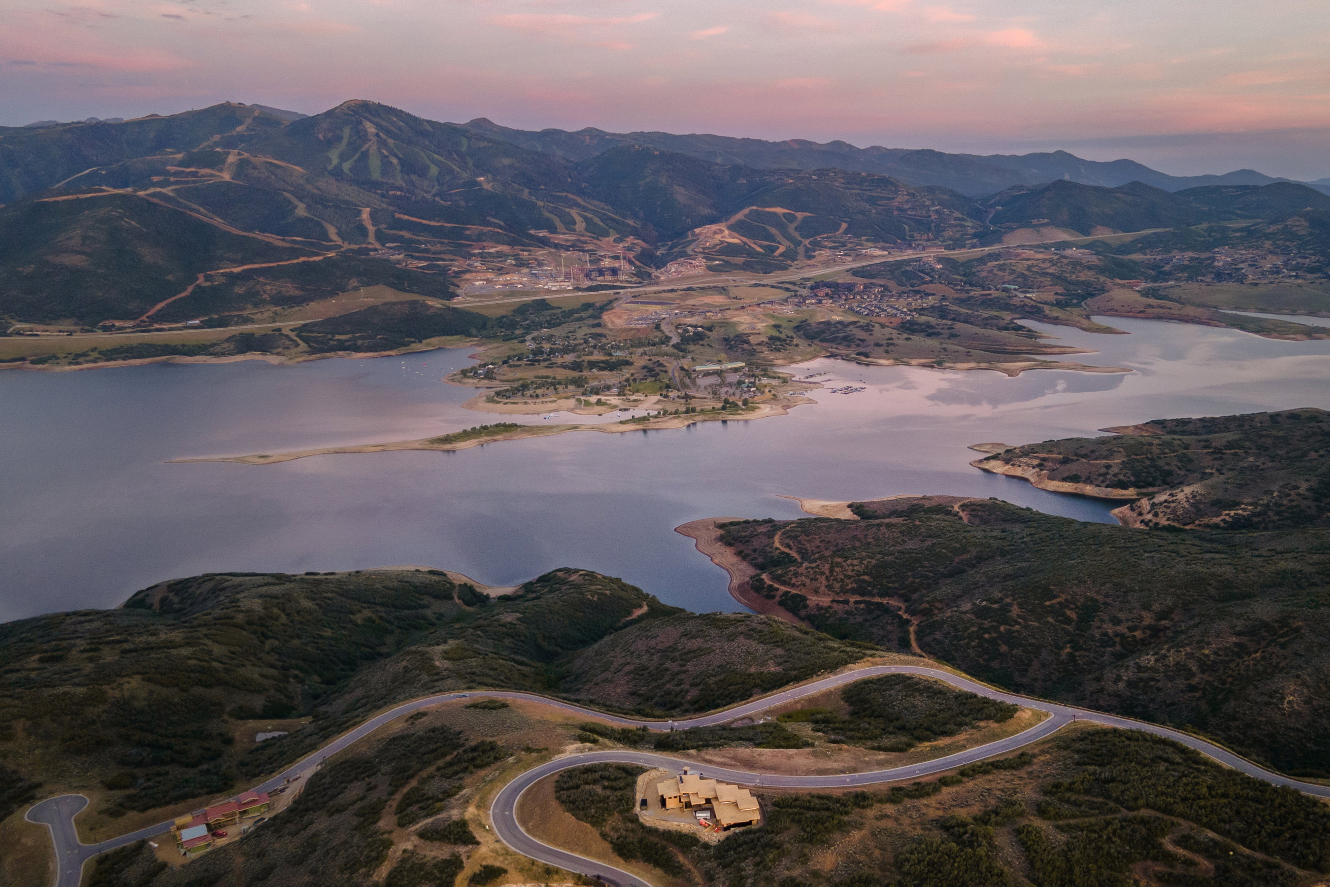 Expansive mountain and lake landscape featuring a winding paved road, surrounding natural vegetation, distant structures, and a prominent body of water