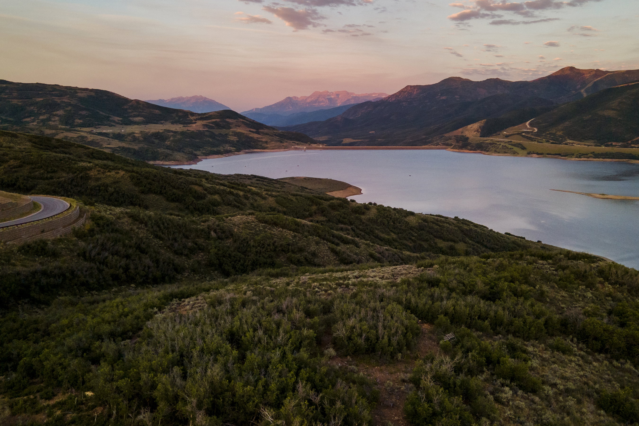 Expansive landscape featuring a large body of water, rolling hills, distant mountains, and winding roadways
