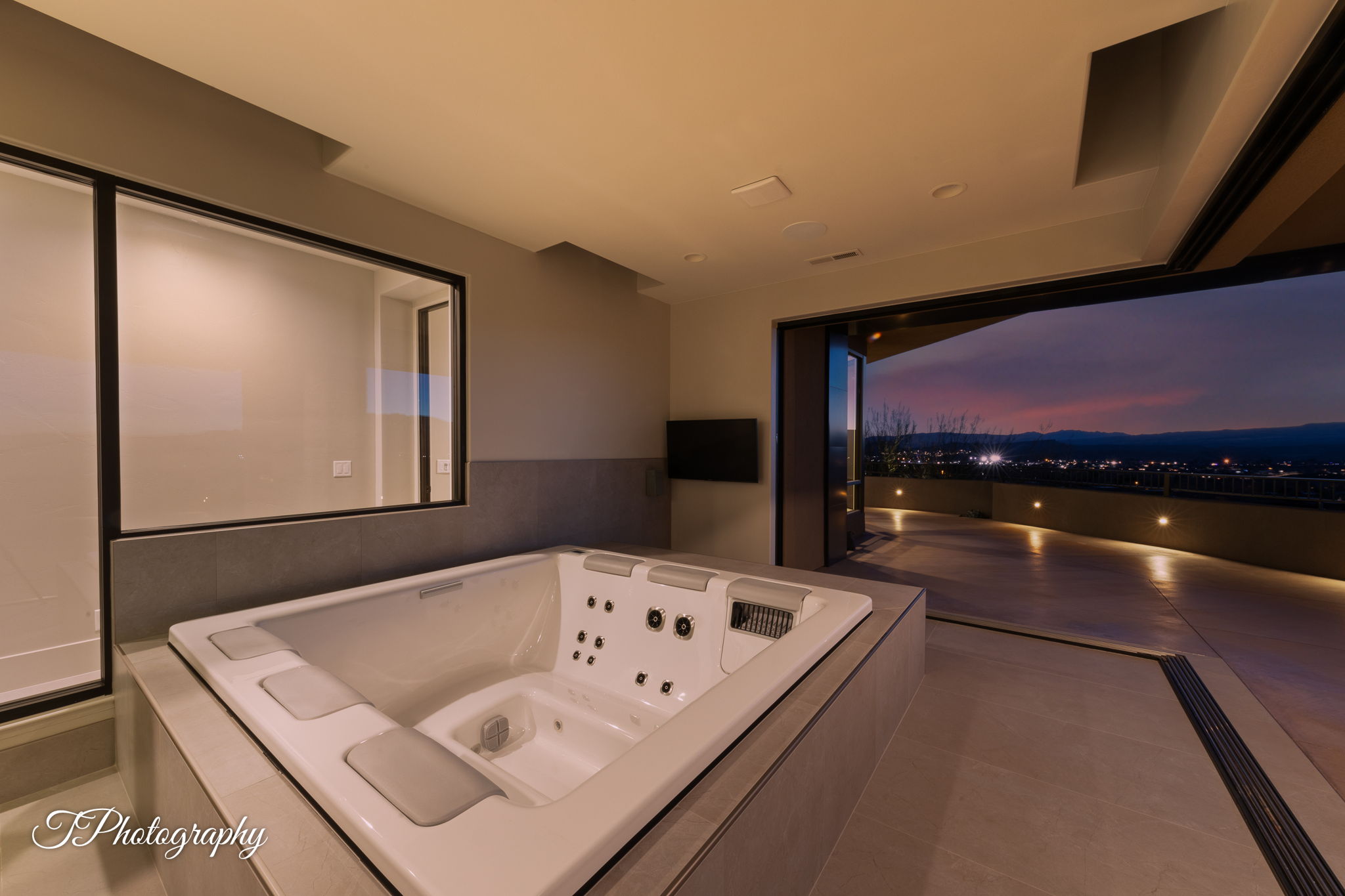 Built-in spa tub with surrounding tilework, expansive retractable glass doors opening to an outdoor patio with recessed lighting, and large interior window framing additional living space