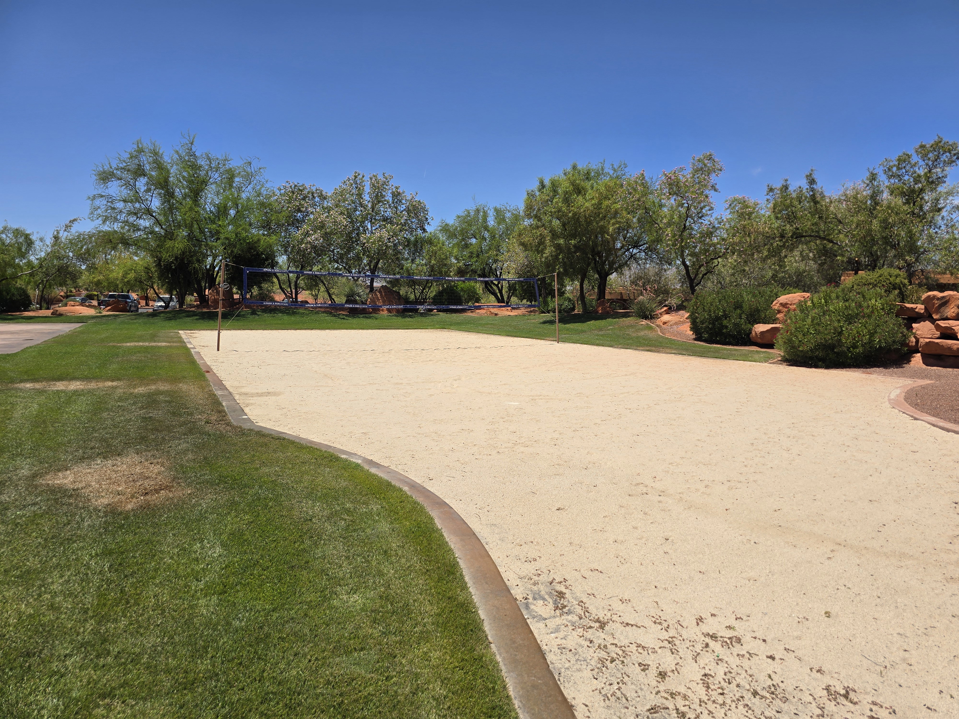 Sand volleyball court featuring a net, surrounded by green lawn and mature trees, with natural rock landscaping