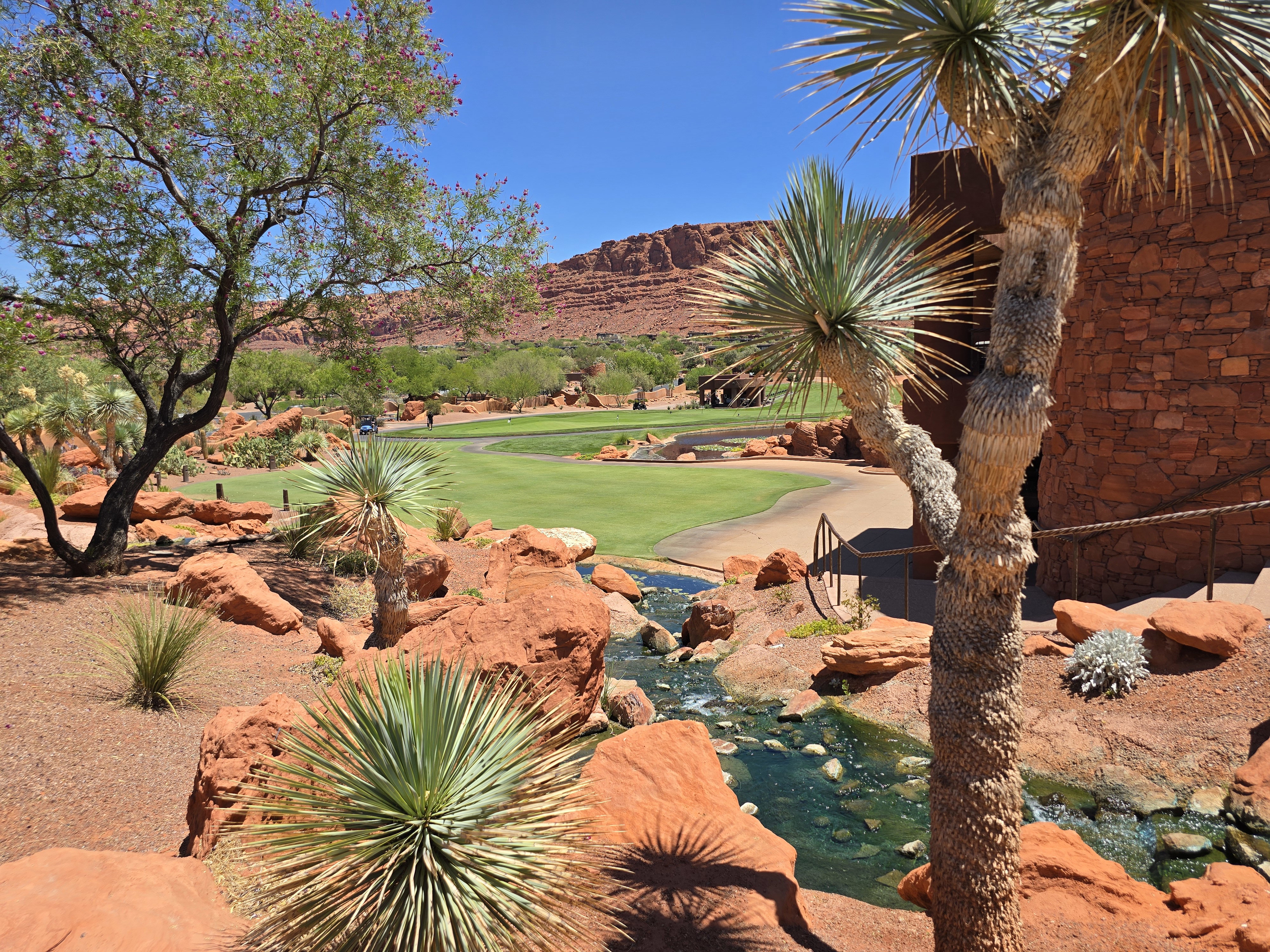 Landscaped exterior featuring a golf course, natural rock formations, desert vegetation, a flowing water feature, and a building with a stone-clad facade