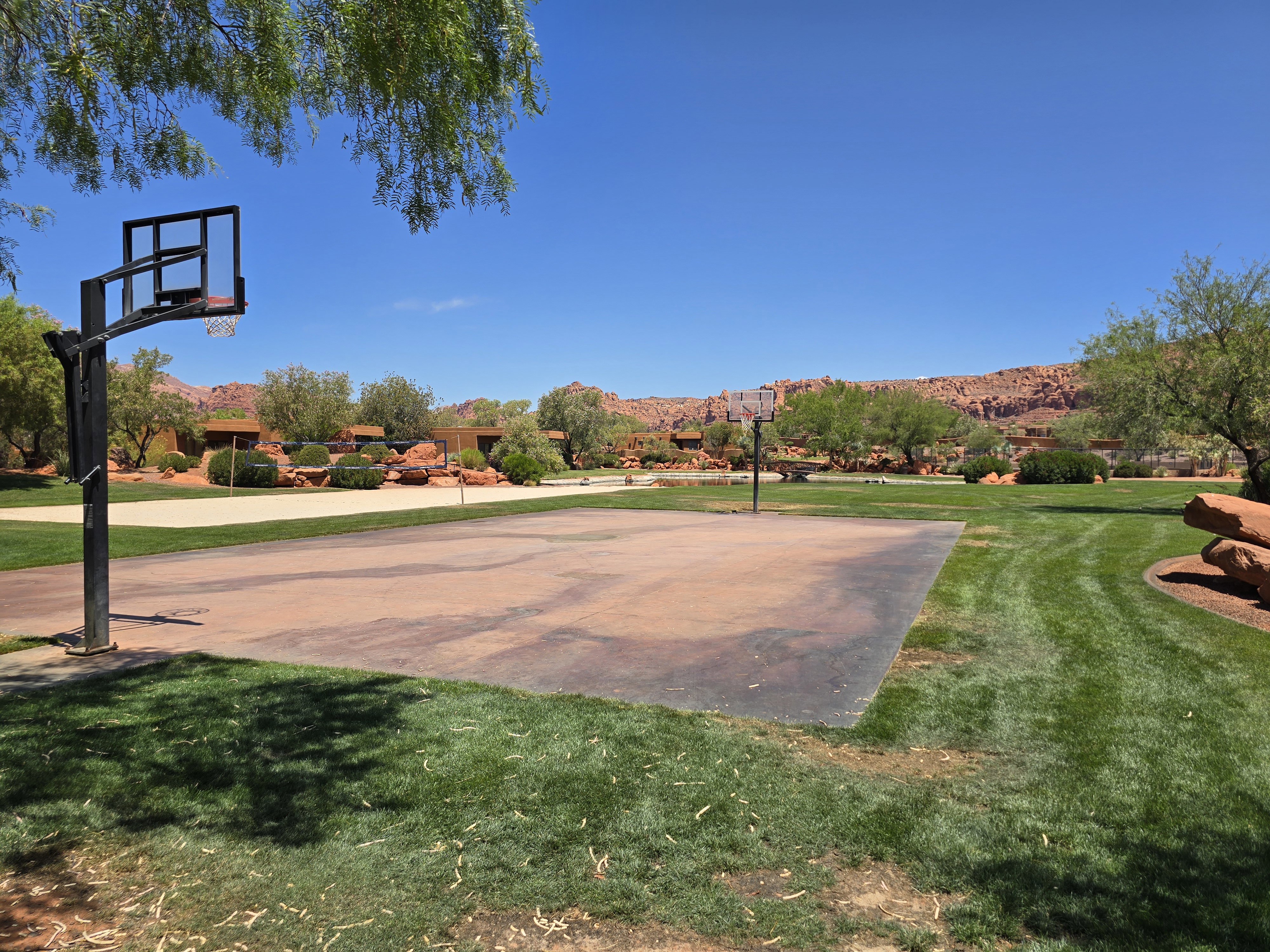 Outdoor sports court featuring a basketball hoop, surrounded by expansive green lawn, mature trees, and distant red rock formations