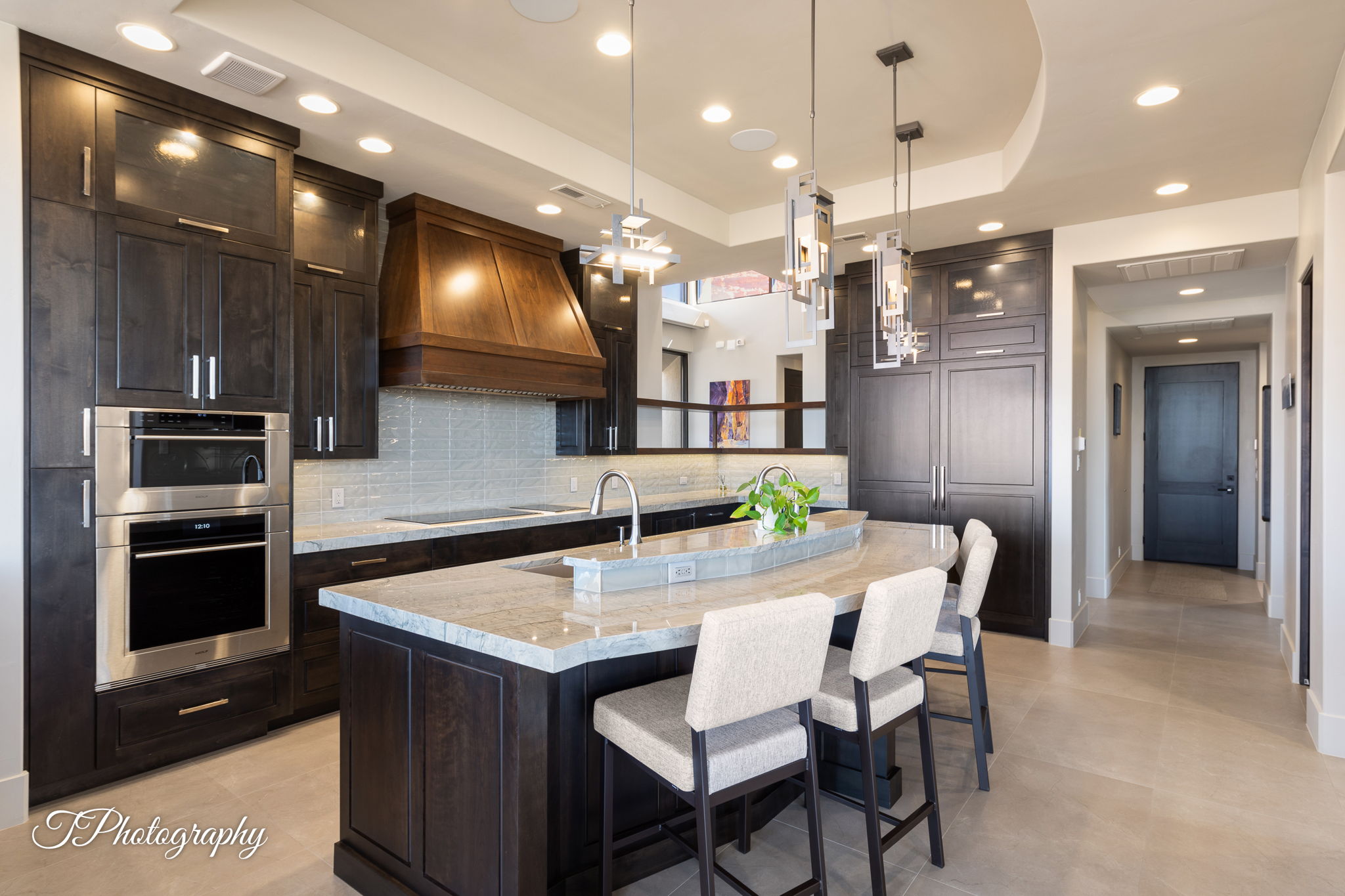 Gourmet kitchen featuring dark wood cabinetry, stainless steel double ovens, a large kitchen island with seating, a custom wood range hood, and pendant lighting