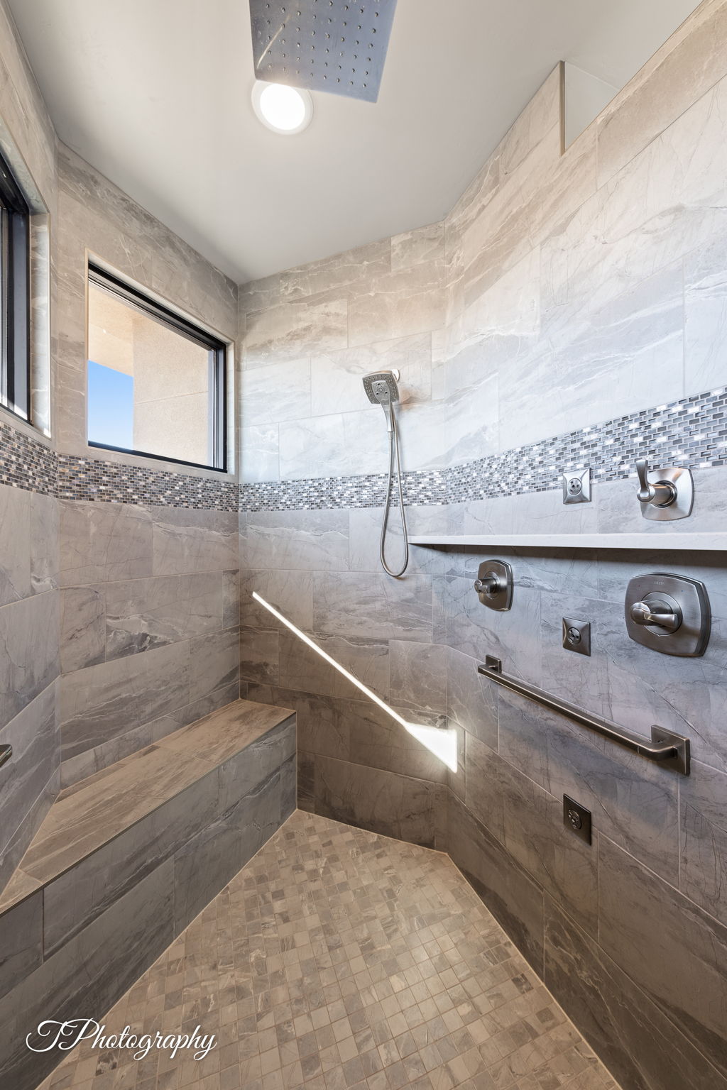 Spacious walk-in shower featuring large format tile walls with a decorative mosaic accent, a built-in bench, dual shower heads, and a window for natural light