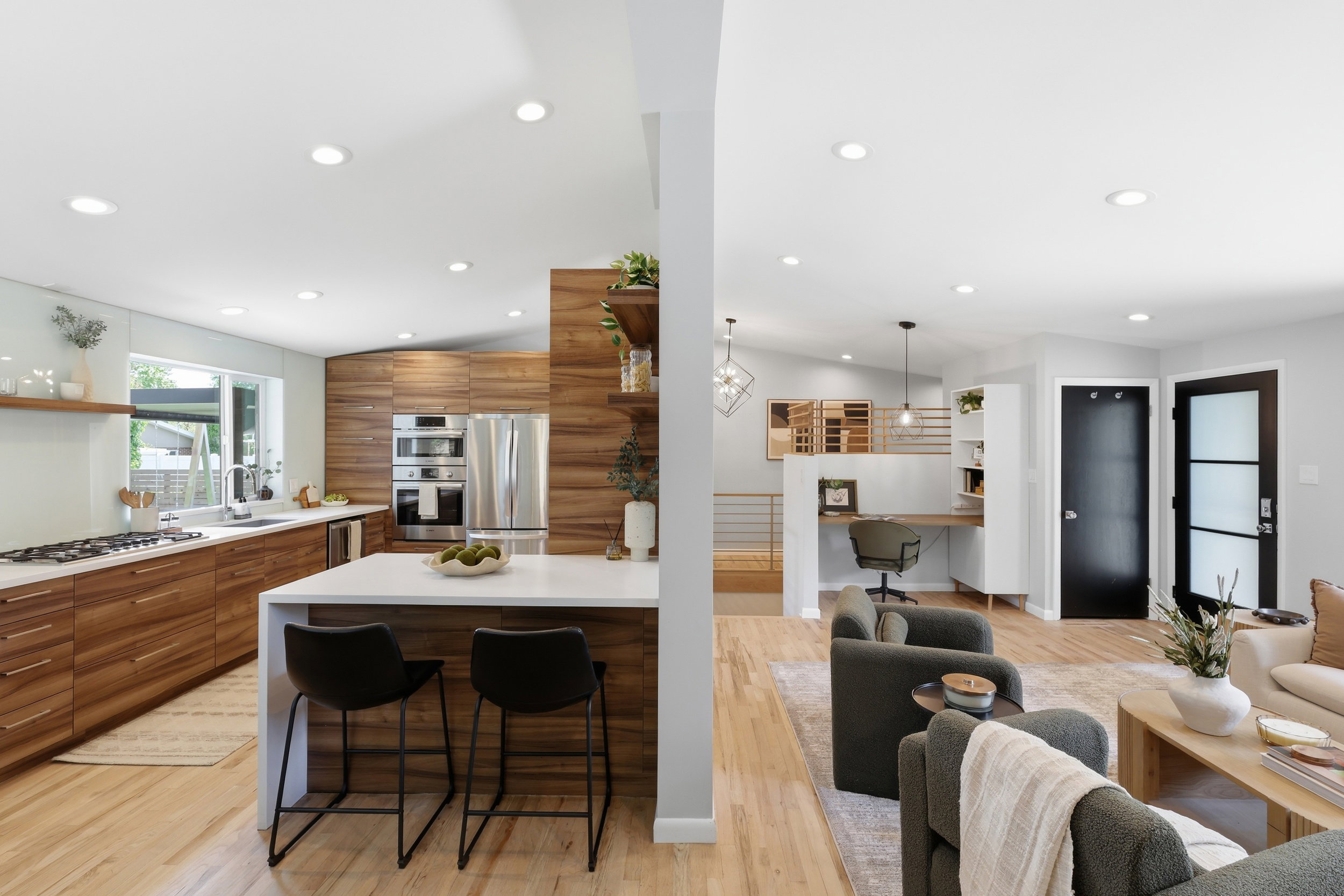 Contemporary open-concept interior featuring light wood-finish flooring, a kitchen with integrated wood cabinetry, a central island with a white countertop, and stainless steel appliances