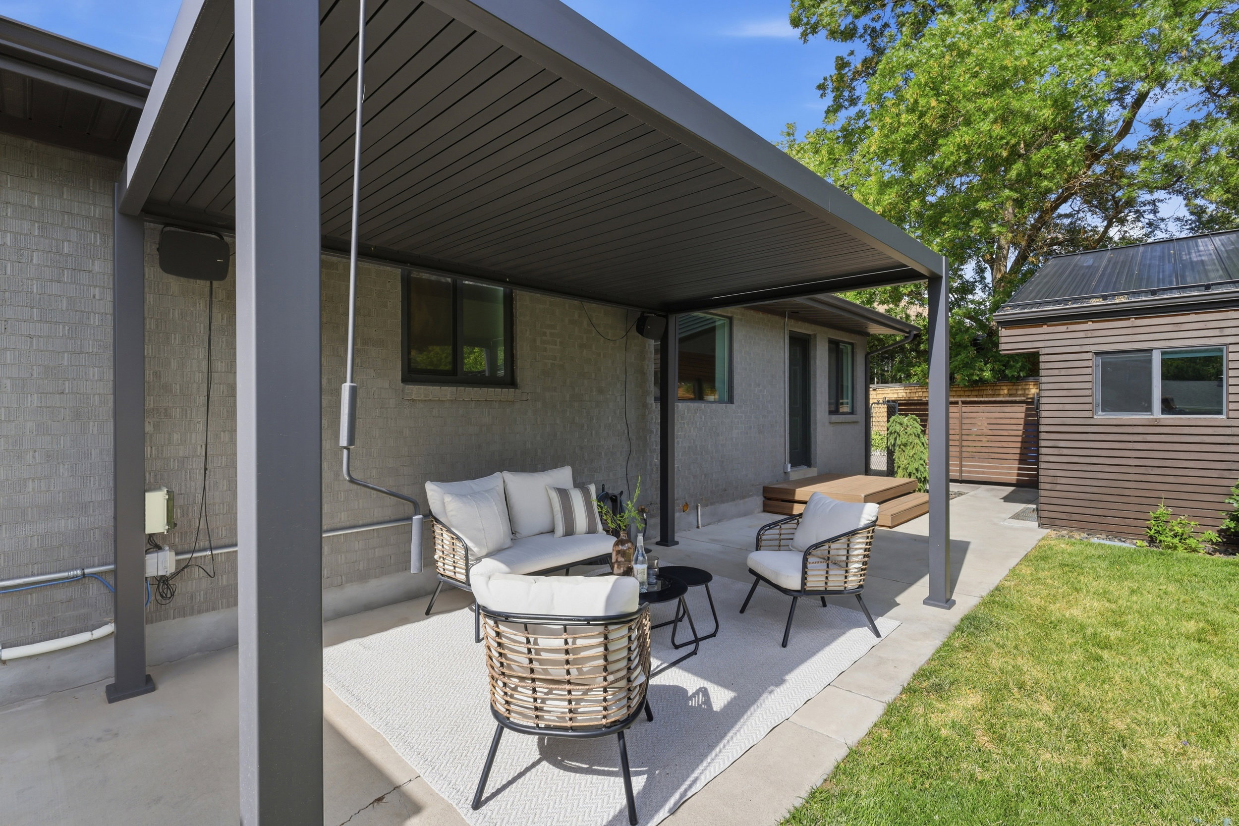 Paved patio area featuring a modern louvered pergola, a wood-finish deck, and a detached structure with horizontal siding and a dark metal roof