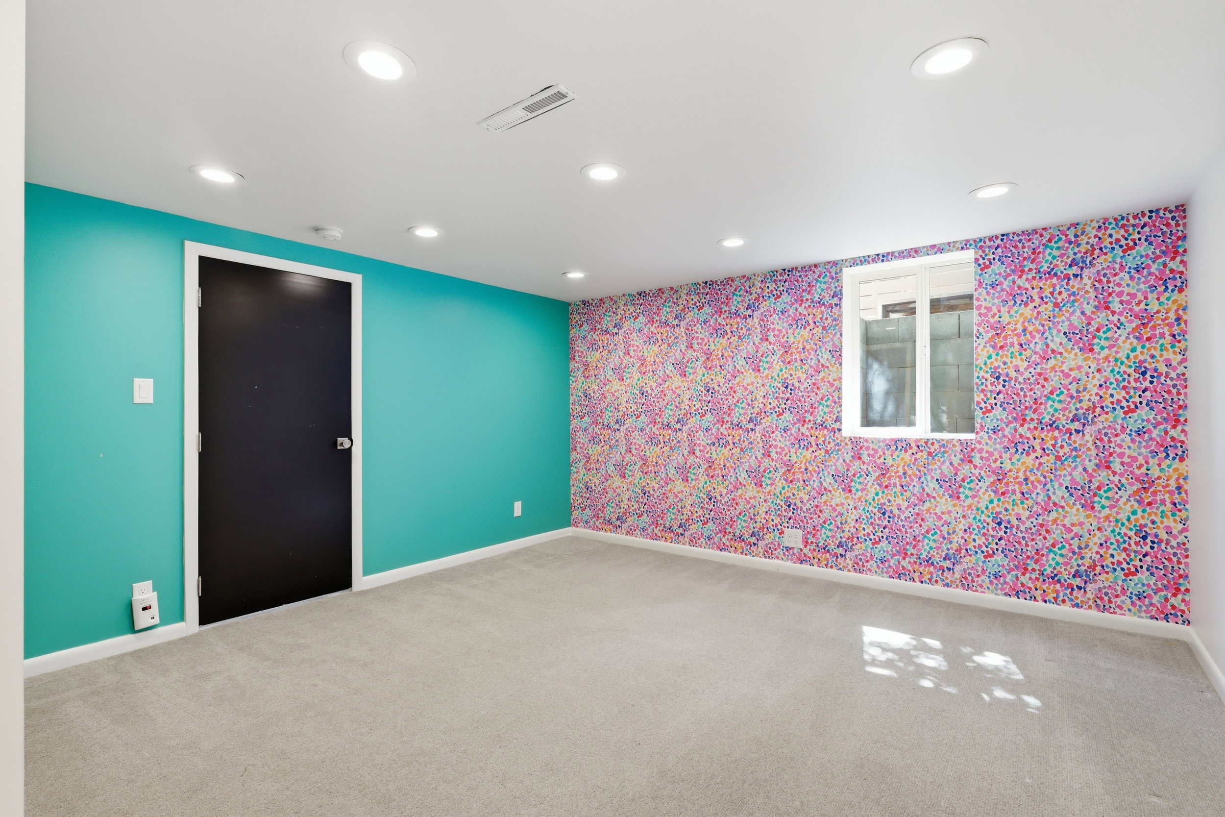 Recessed ceiling lighting illuminates a room featuring textured gray carpeting, a bright teal painted wall, and a multicolor patterned accent wall