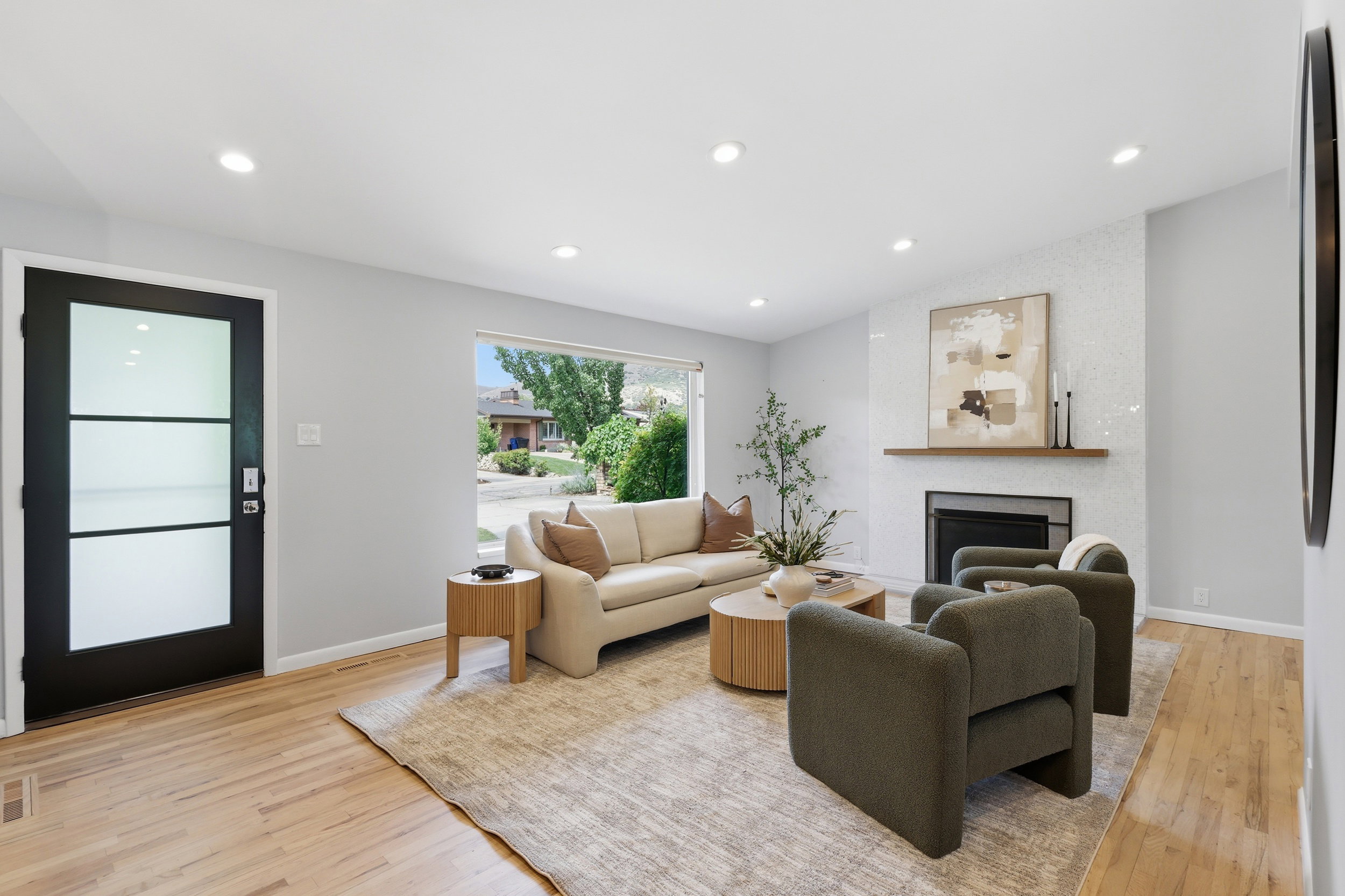 Bright living space featuring wood-finish flooring, a contemporary fireplace with a decorative tile surround, a dark framed entry door with frosted glass panels, recessed lighting, and a large window providing exterior views