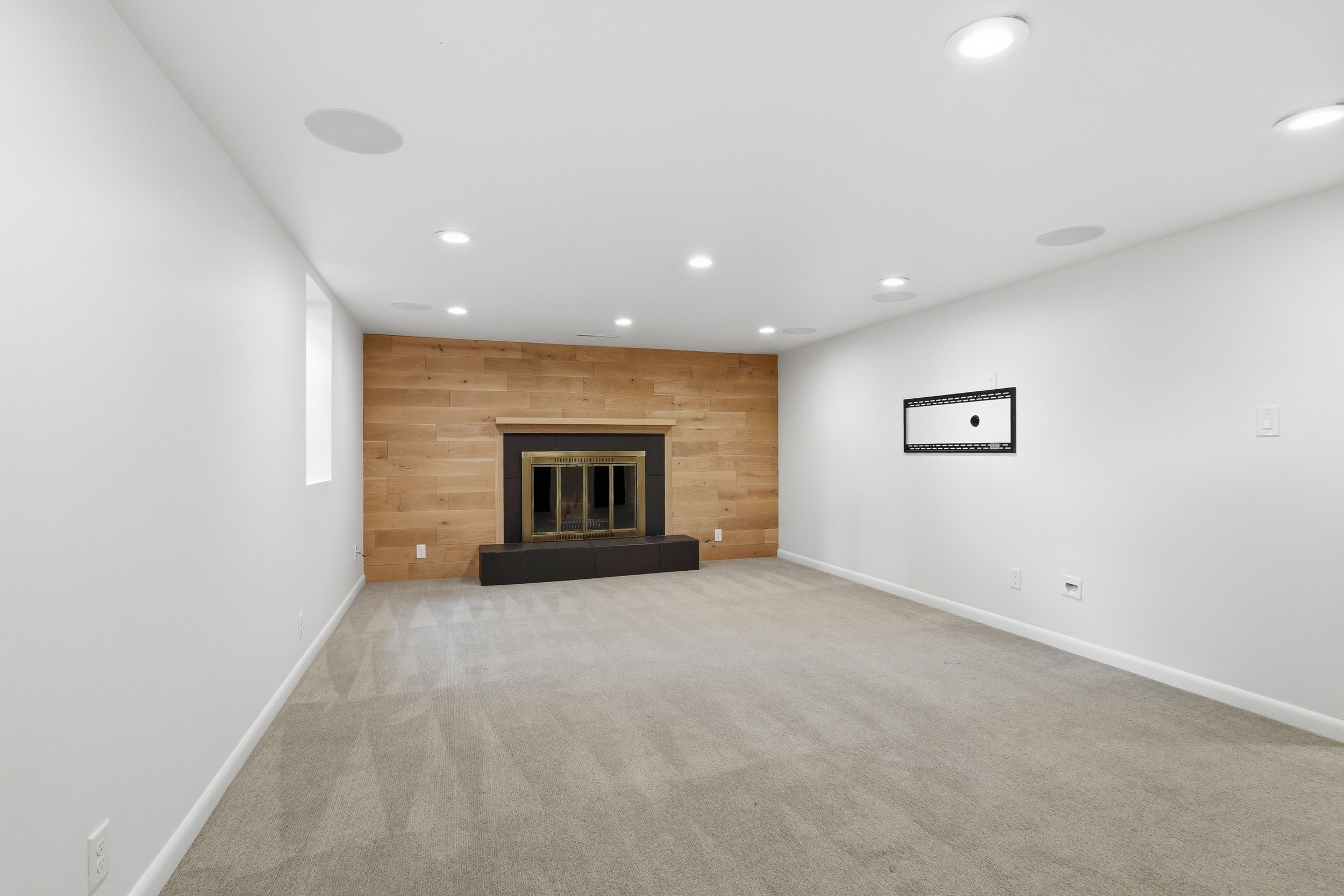 Spacious room featuring a wood-paneled accent wall with a fireplace, recessed lighting, ceiling-mounted speakers, and light-colored carpet flooring