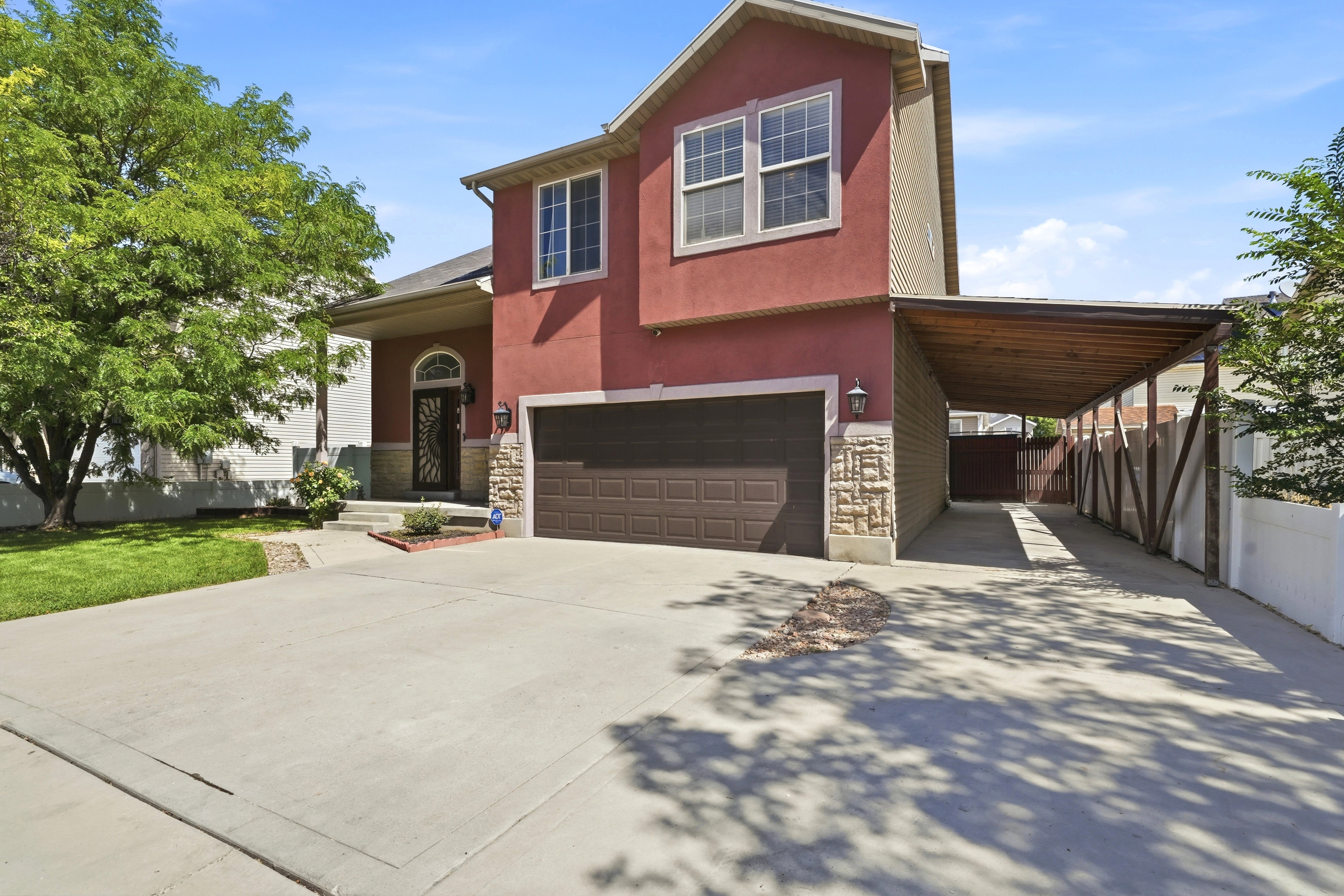 Two-story residence featuring a red stucco exterior with stone accents, an attached two-car garage, a covered carport, a concrete driveway, and a manicured lawn