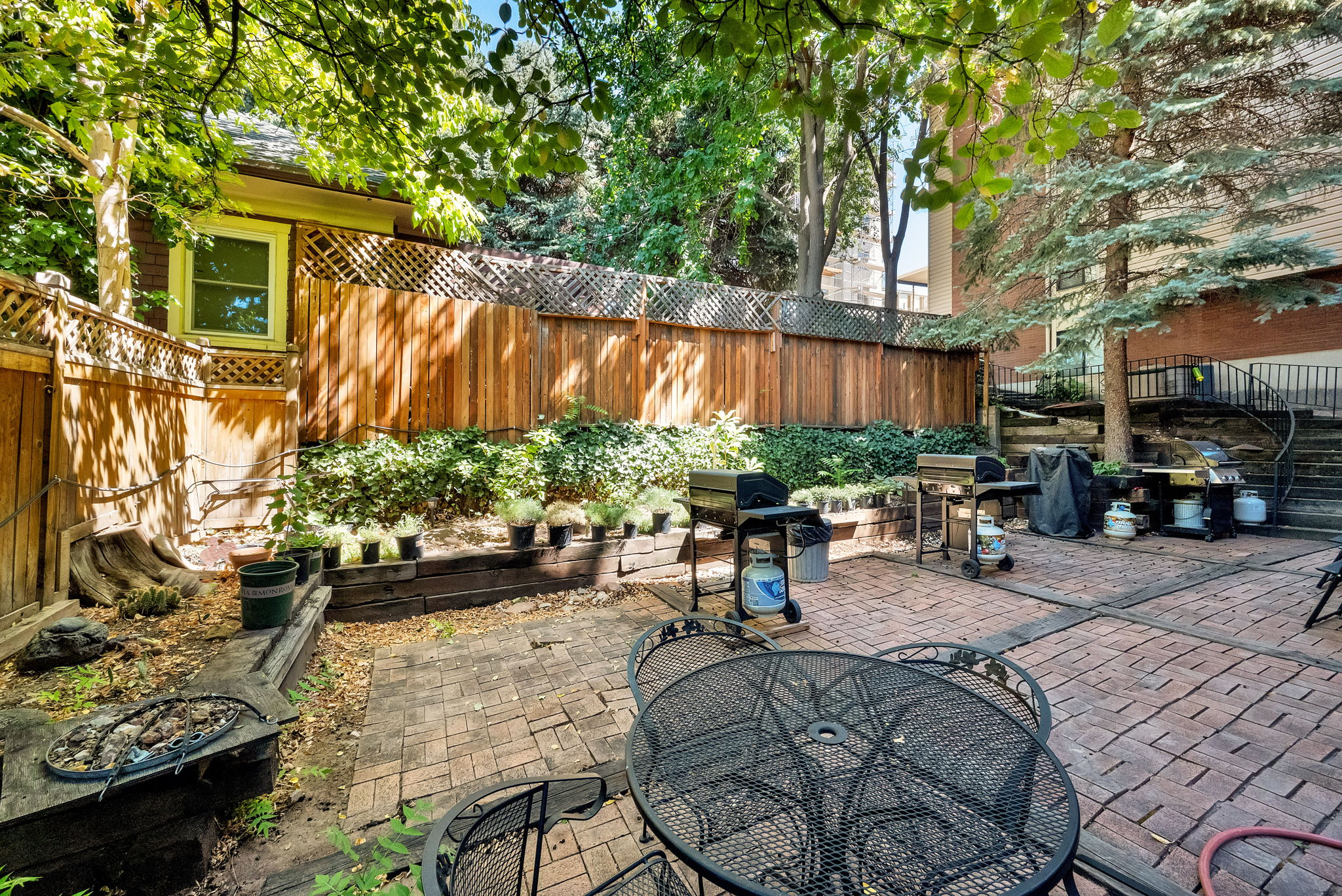 Expansive brick paver patio featuring a perimeter wood fence, tiered landscaping, mature trees, and a visible building exterior